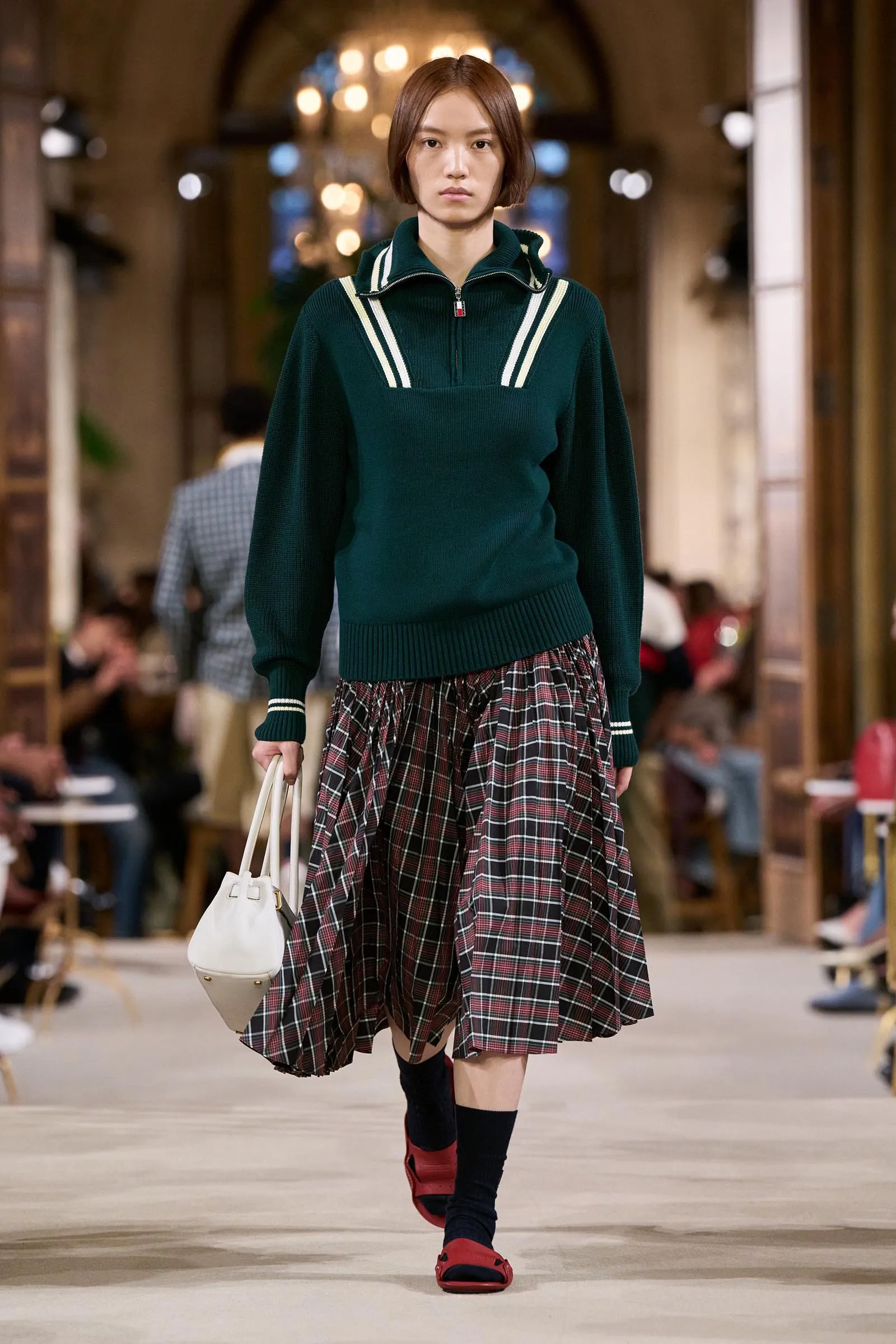 Model walking a fashion runway in a dark green sweater with white striped accents and a plaid midi skirt, carrying a white handbag.