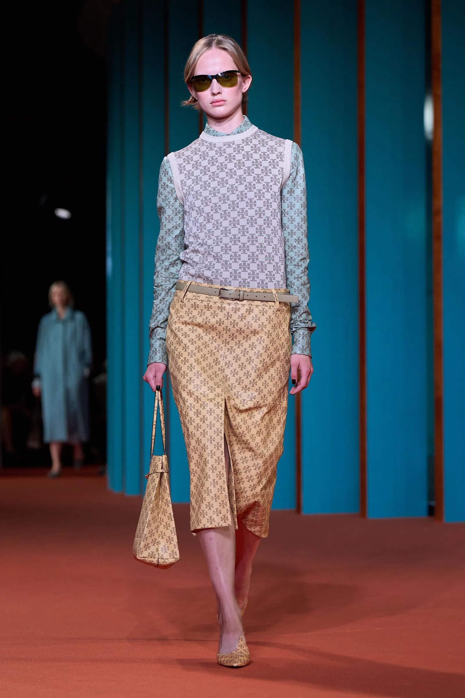 Model walking a fashion runway in a beige patterned jumpsuit with a pale gray sleeveless knit top, tan belt, and sunglasses against teal panels.