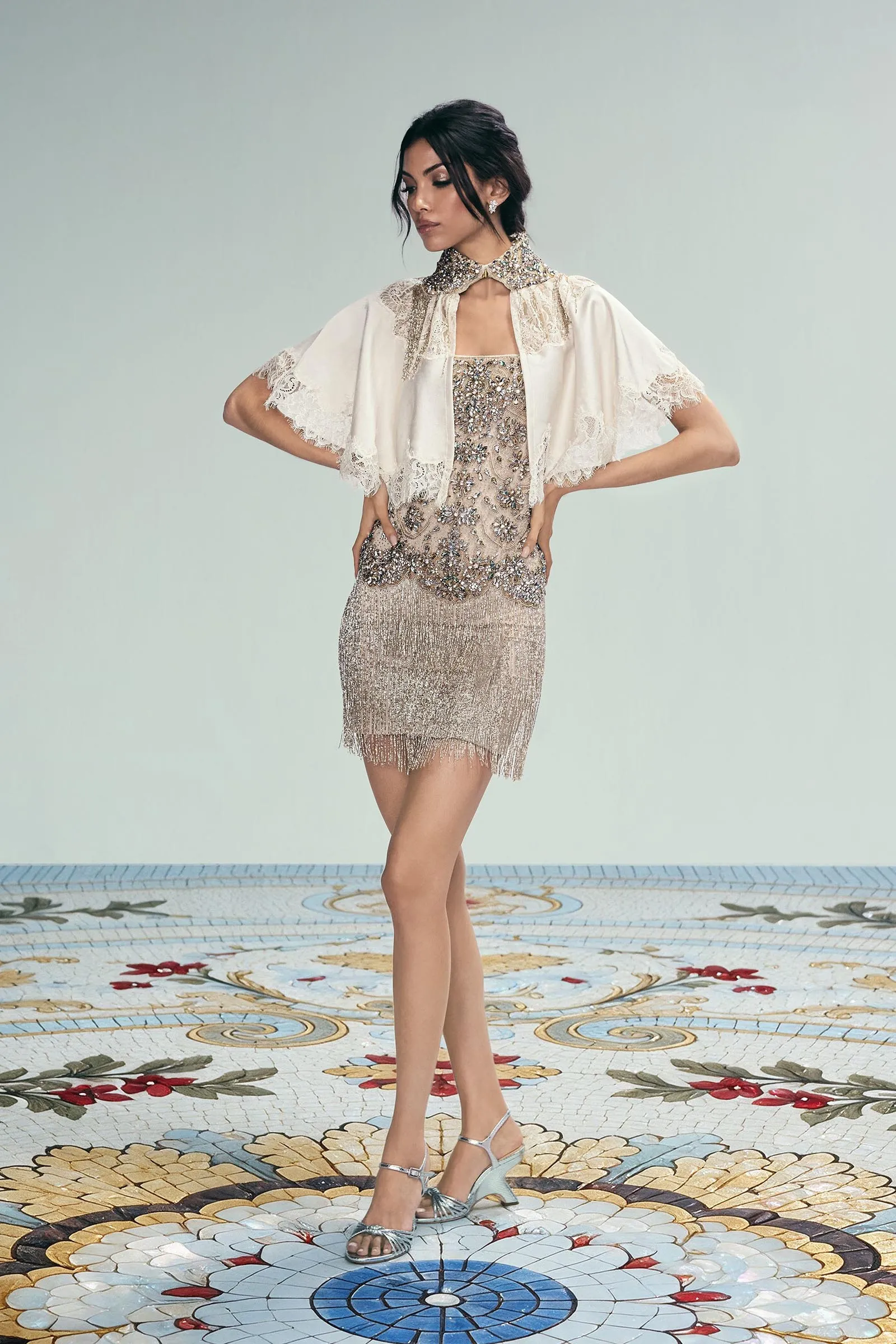 Model wearing a champagne beaded mini dress with a jeweled collar and lace cape, posing on a decorative mosaic floor.