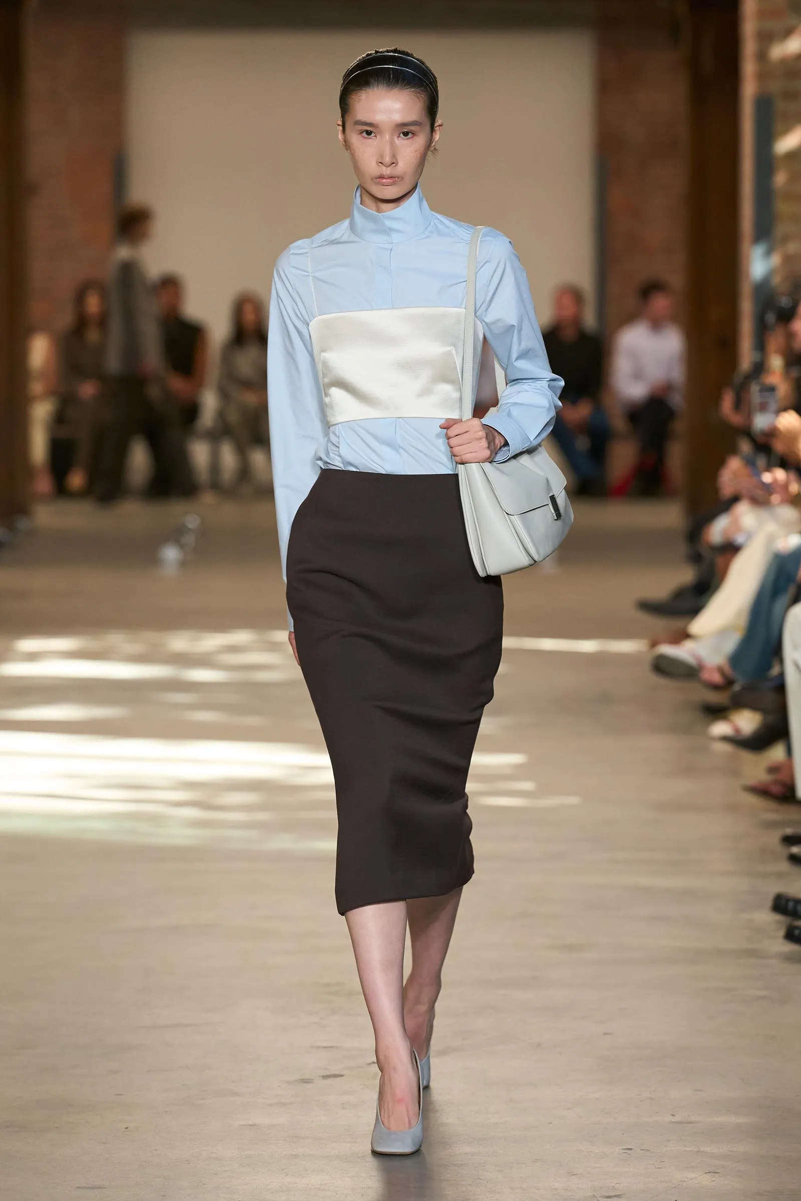 Model walking a runway in a pale blue high-neck blouse with a white sash, paired with a black pencil skirt and light gray handbag.