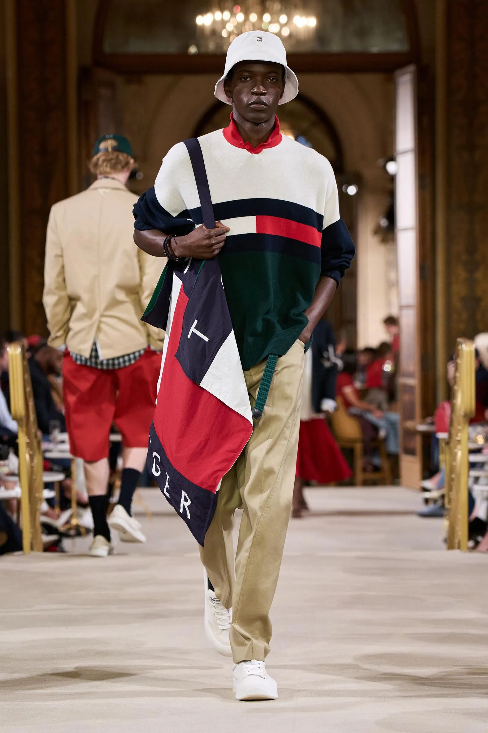 Male model walks a fashion runway in a color-block sweater, beige pants, white bucket hat, and a large flag-pattern tote over his shoulder in a grand venue.
