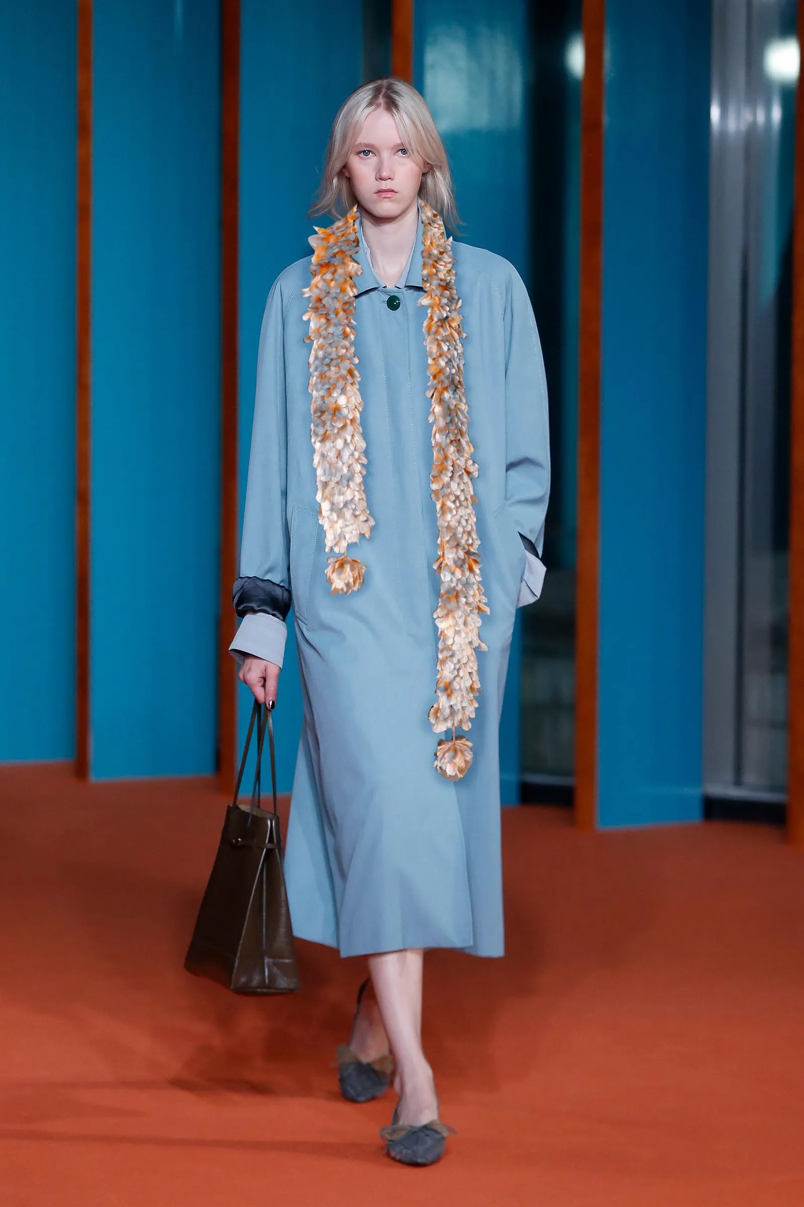 Model on a runway wearing a light blue coat and a long shell-adorned scarf, carrying a brown handbag against a blue backdrop.