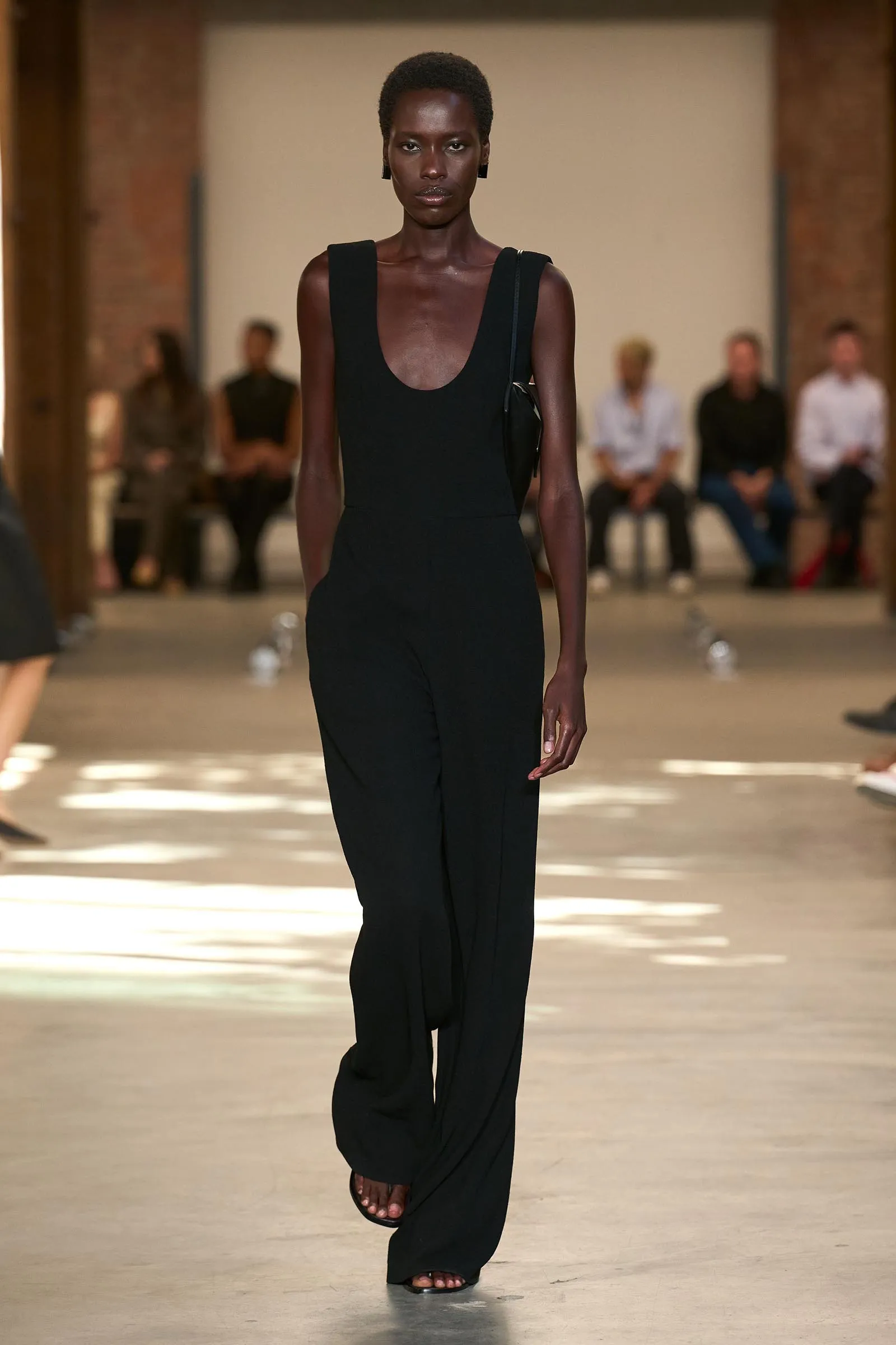 Model walks the runway in a black sleeveless jumpsuit with a deep scoop neckline and a small black shoulder bag.