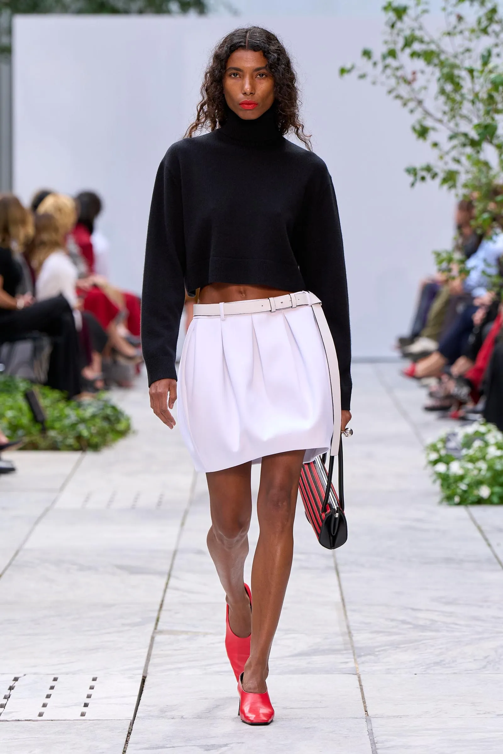 Model walking runway in a black cropped turtleneck and white belted pleated skirt, wearing red shoes; audience blurred in background.