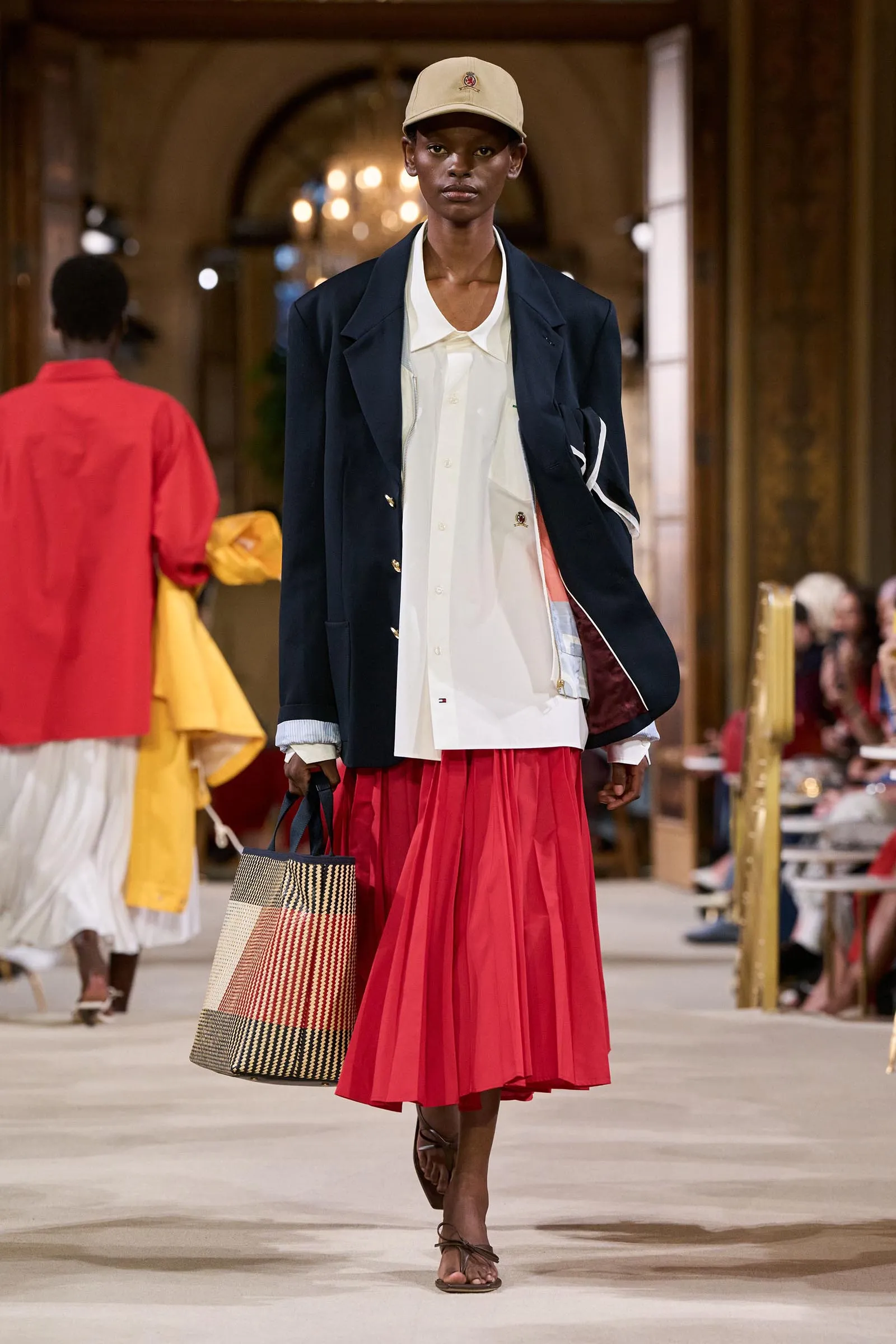 Model walking a fashion runway in a navy blazer, white shirt, and a bright red pleated skirt, with a beige cap and a large plaid tote bag.