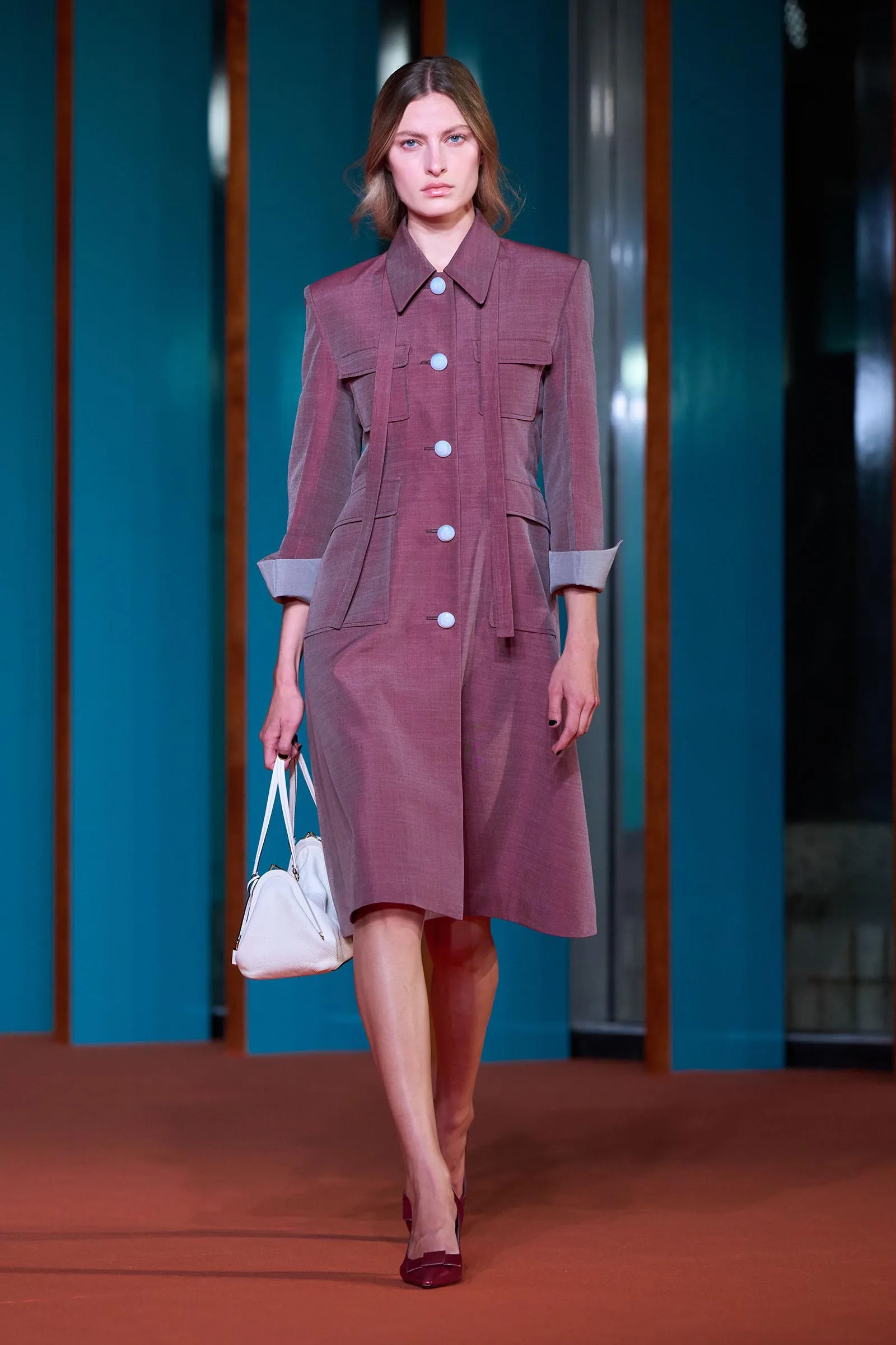 Model walking a runway in a dusty-rose dress with large light-blue buttons and a white handbag on her left side.