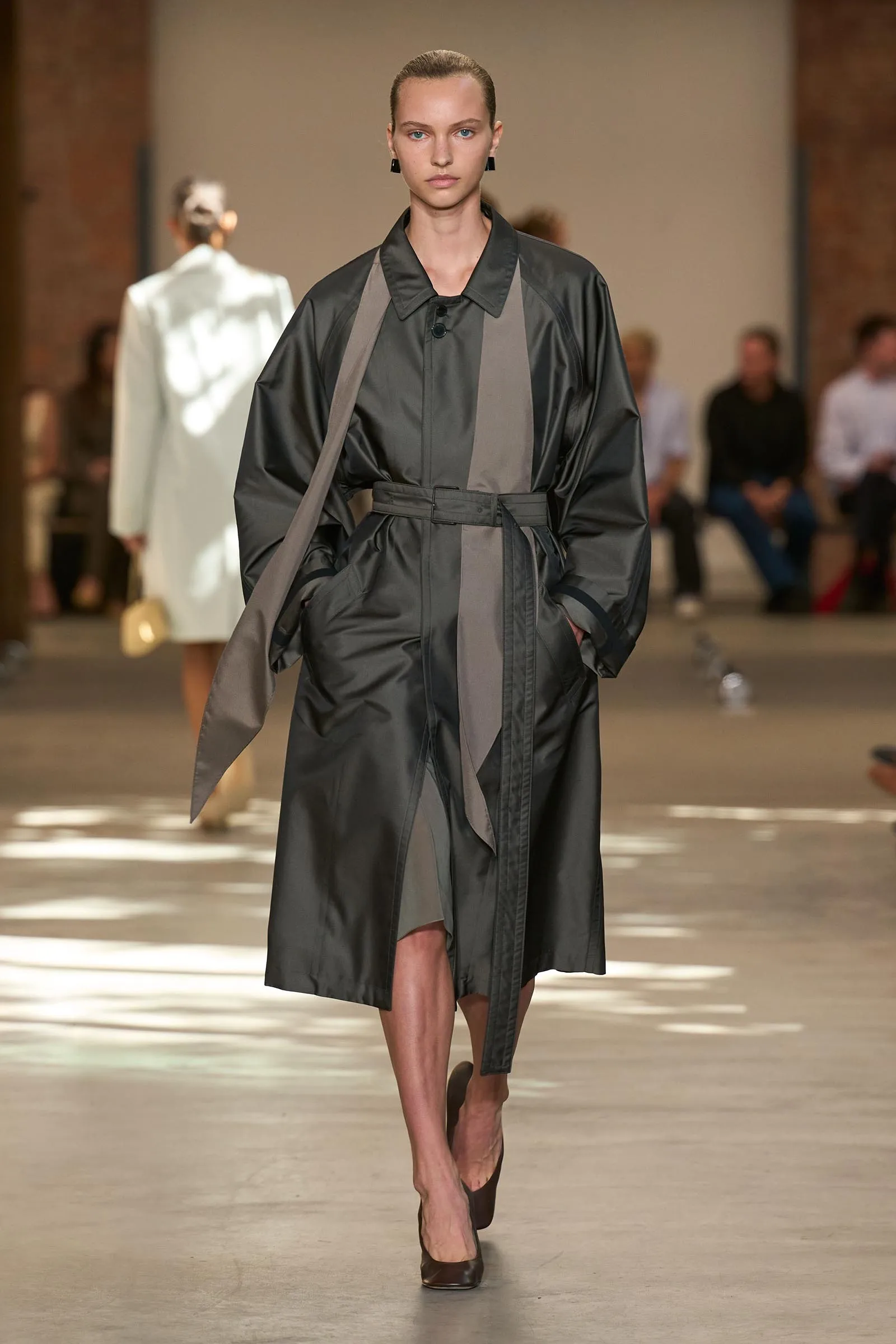 Model walking a runway in a dark gray trench coat with a belt and lighter gray panels, wearing high heels.