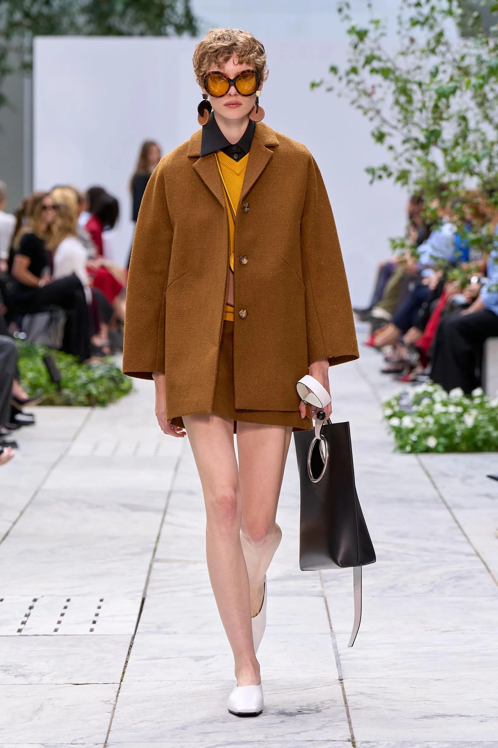 Fashion model walking a runway in a brown oversized coat over a mustard sweater, with bold orange round sunglasses and large circular earrings, carrying a black bag.
