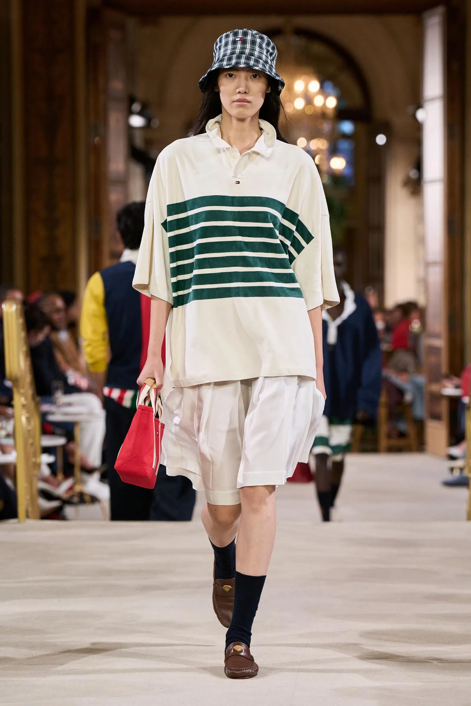 Model walking a fashion show in an oversized cream top with green horizontal stripes, white shorts, a plaid bucket hat, and brown loafers; carrying a red bag.