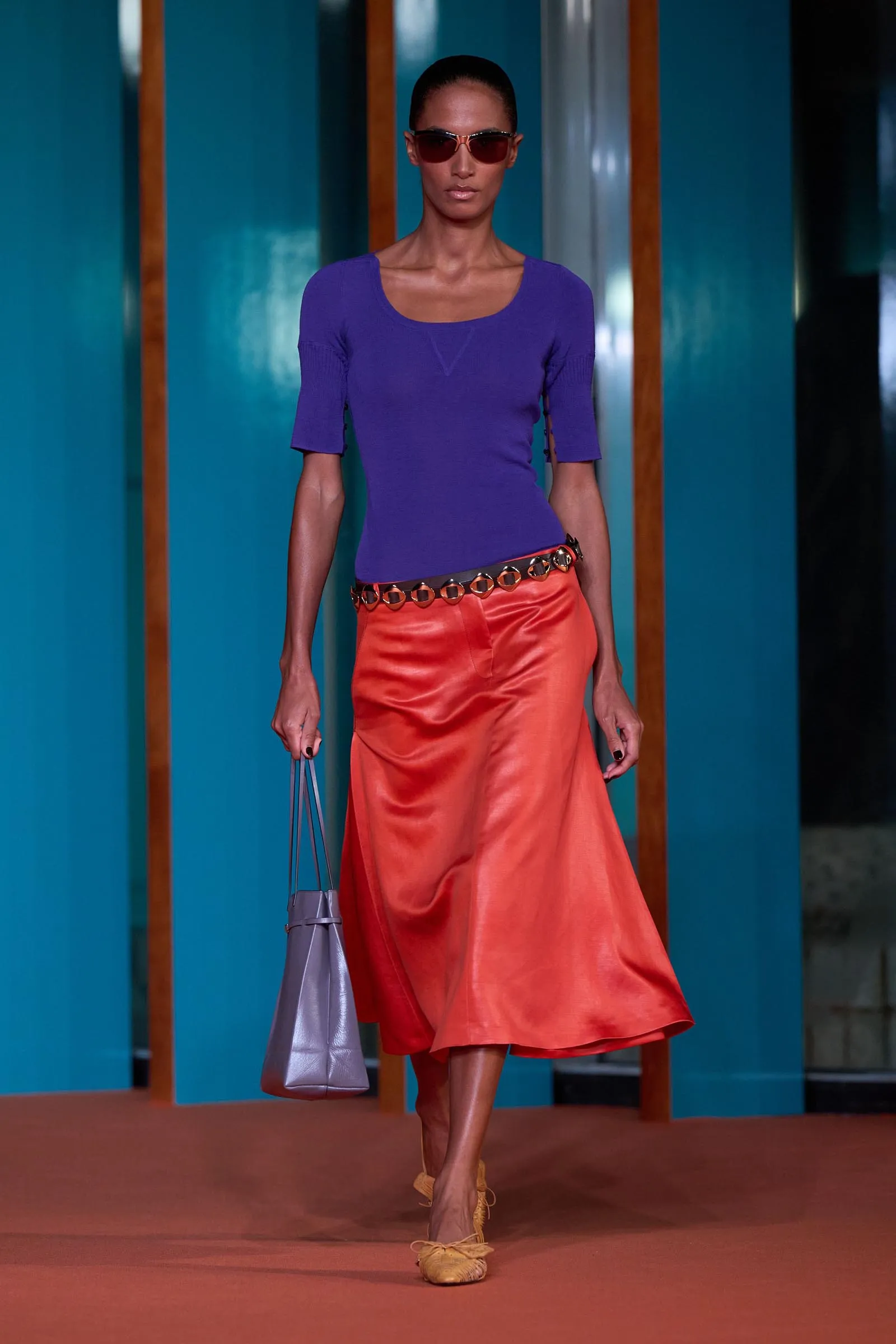 Model walking a fashion runway in a purple top and orange satin skirt, wearing sunglasses and carrying a lavender handbag.