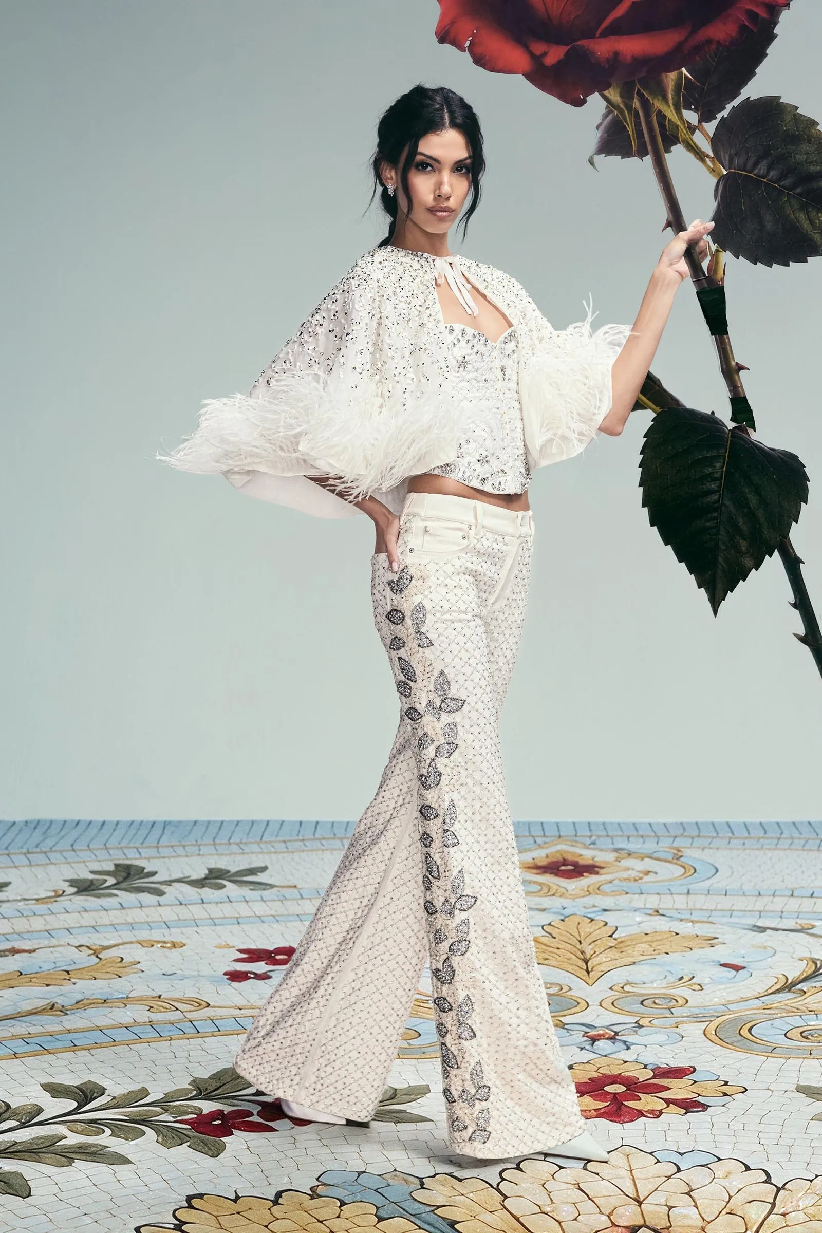 Fashion model in a sparkling white, beaded top and patterned pants, posing with a giant red rose against a pale teal backdrop.