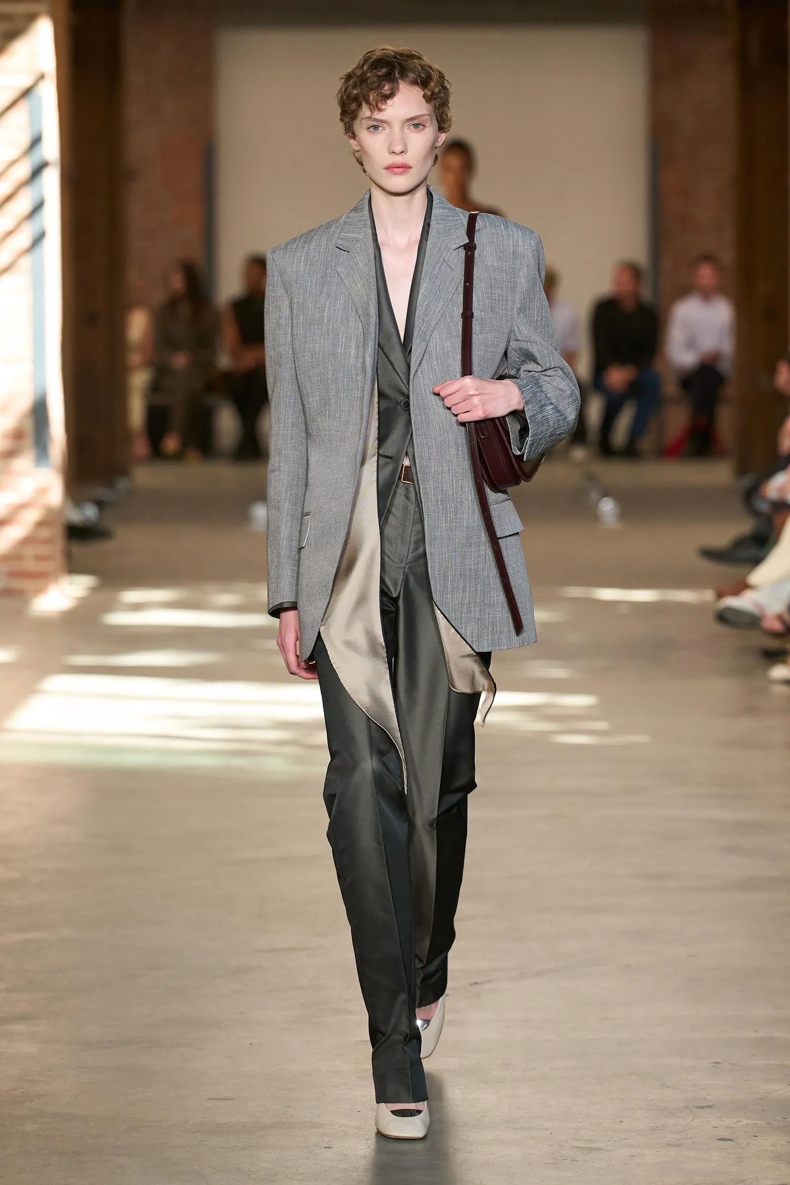 Model walking a fashion runway in a light gray oversized blazer with dark trousers and a burgundy shoulder bag.