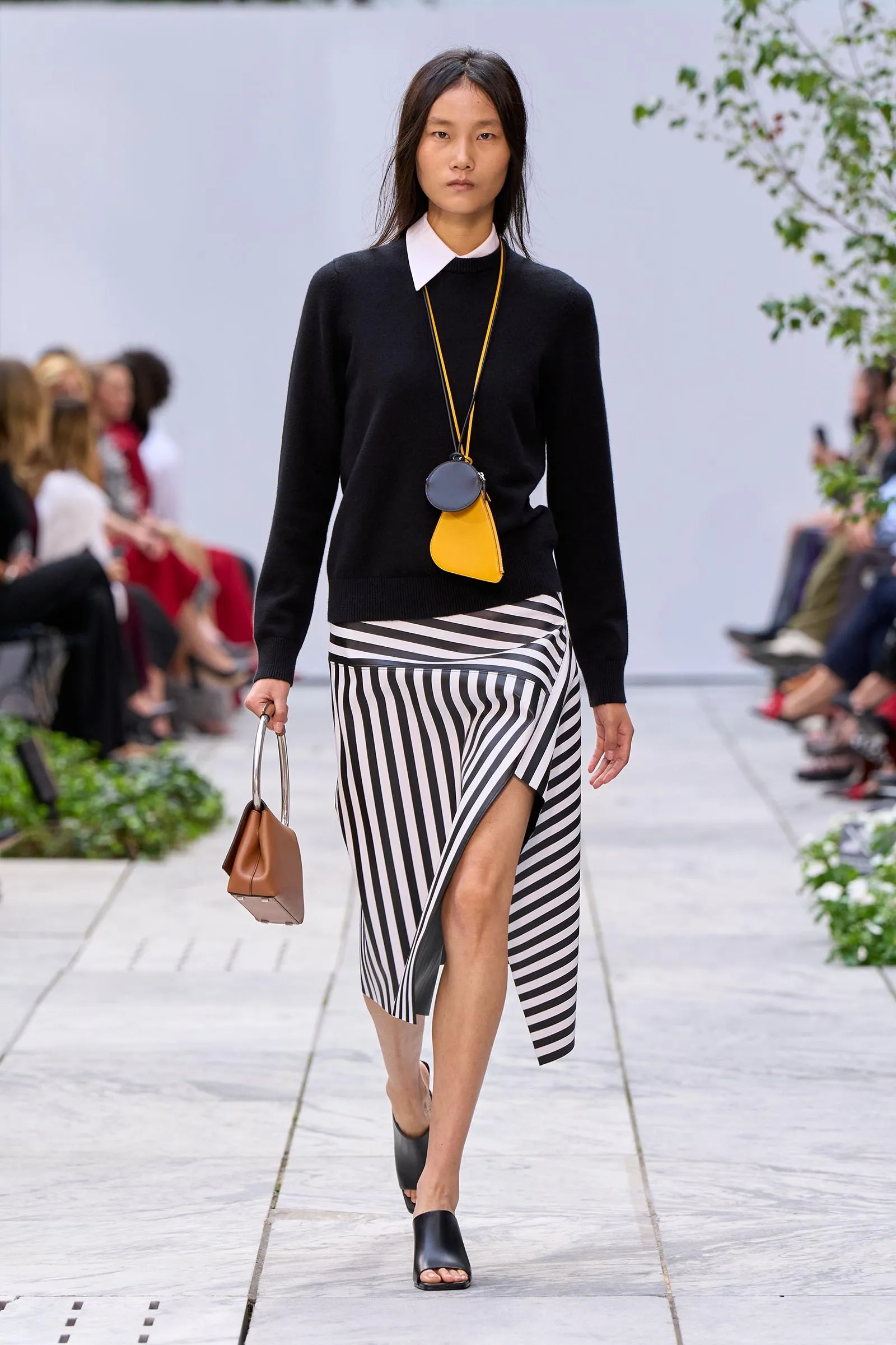 Model walking runway wearing a black sweater with white collar and a black-and-white diagonal-striped skirt, accessorized with a yellow crossbody bag and round dark coin purse; holding a brown handbag.