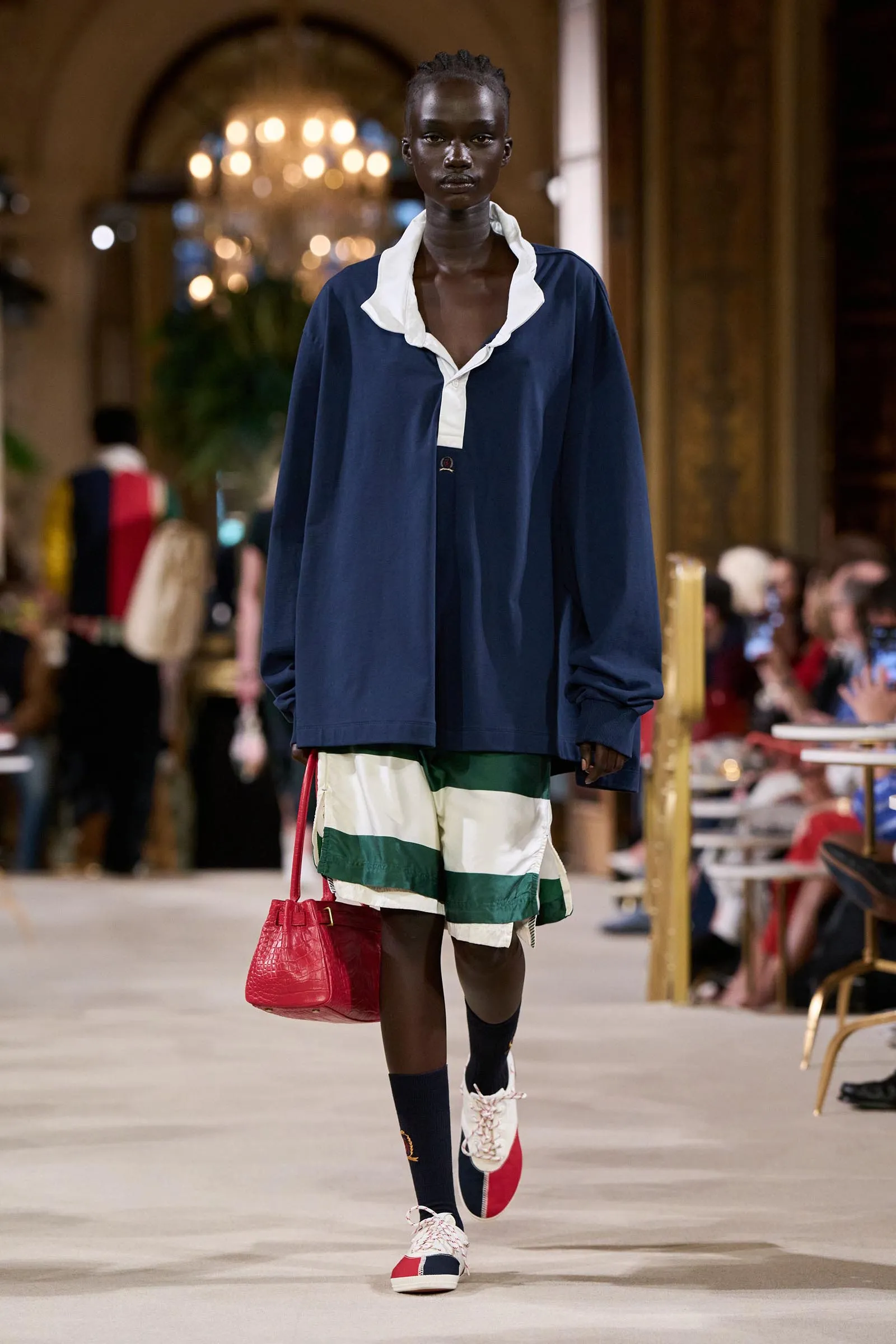 Model walking runway in a navy top with white collar, striped green and white shorts, and red handbag in a grand venue with chandeliers.