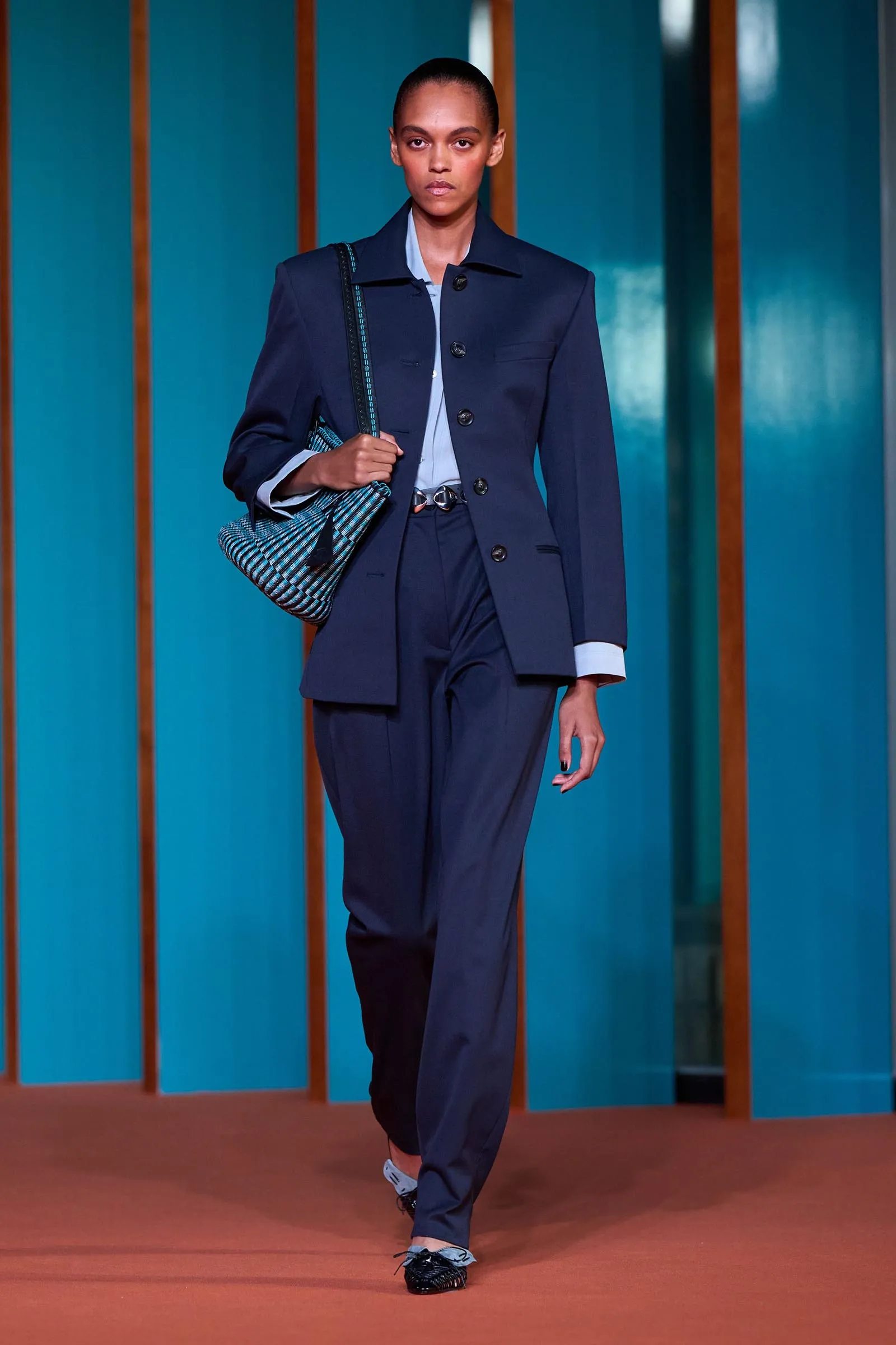 Female model walking a runway in a navy suit, carrying a blue striped bag against a teal panel backdrop.