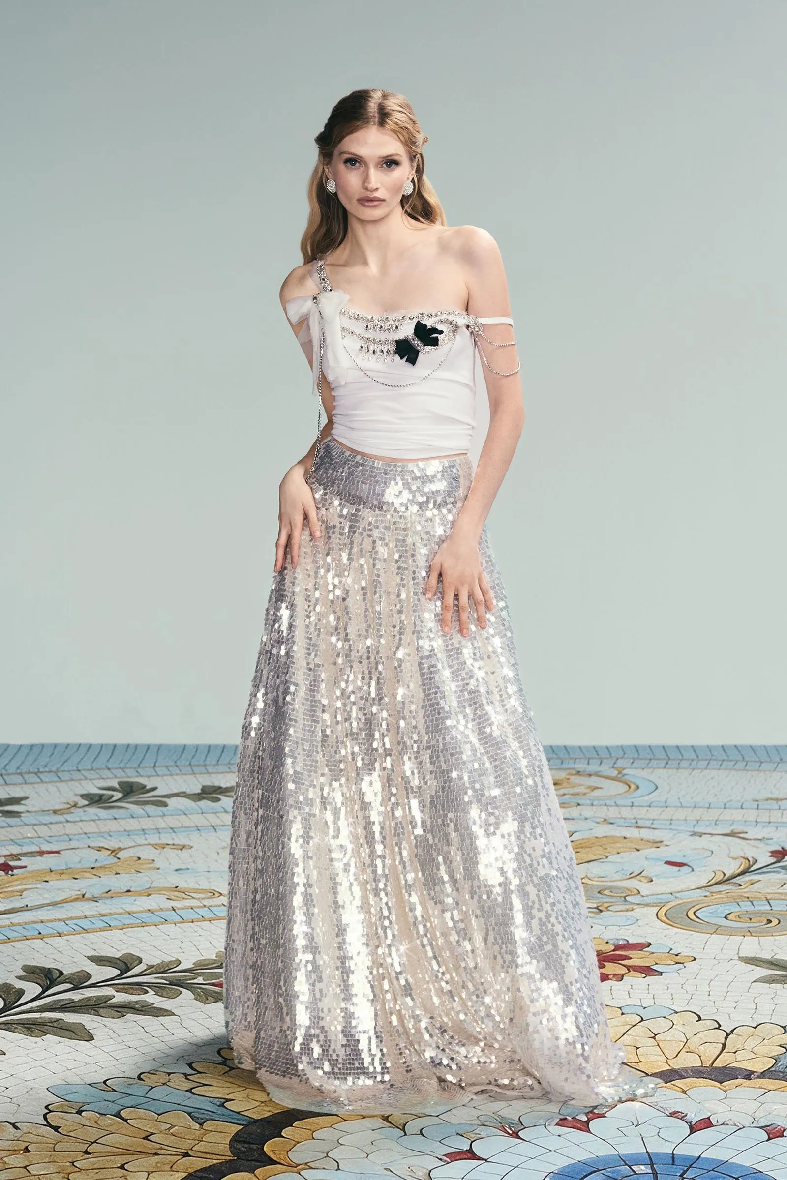 Model wearing a white off-shoulder top with a silver sequined skirt, posing against a pale background.