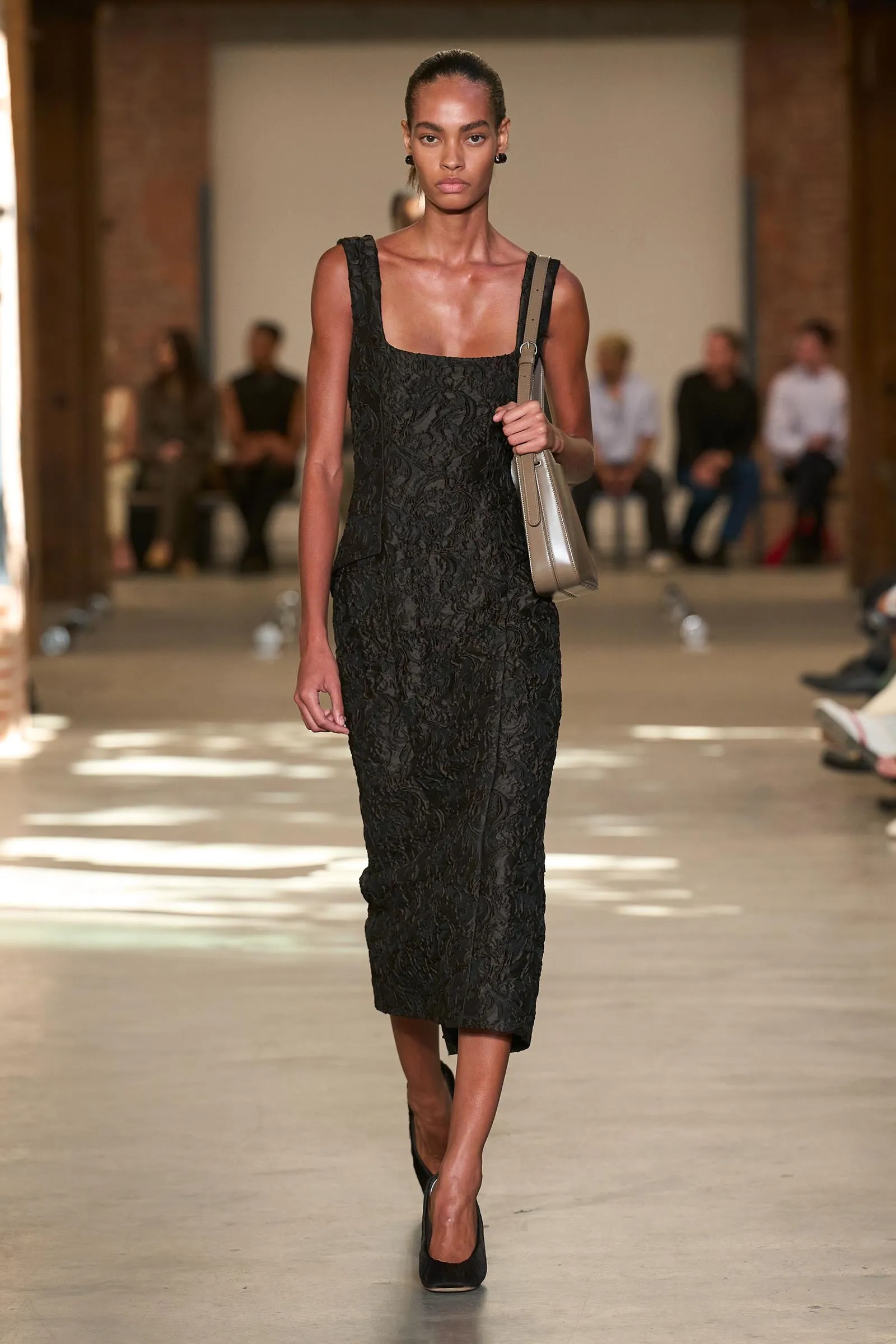 Model walking the runway in a black lace sleeveless midi dress, carrying a beige shoulder bag.