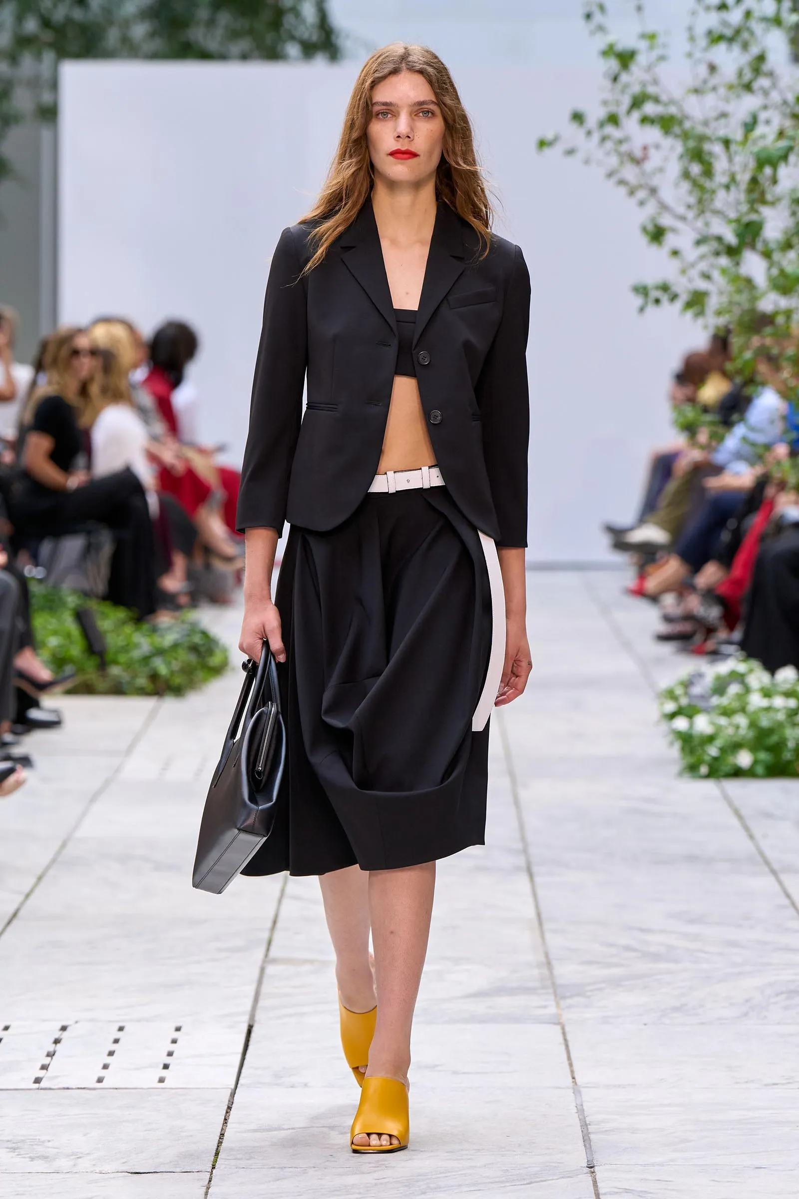 Model walking a runway in a black blazer over a cropped top, a draped black skirt with a white belt, and bright yellow open-toe sandals, carrying a gray bag.