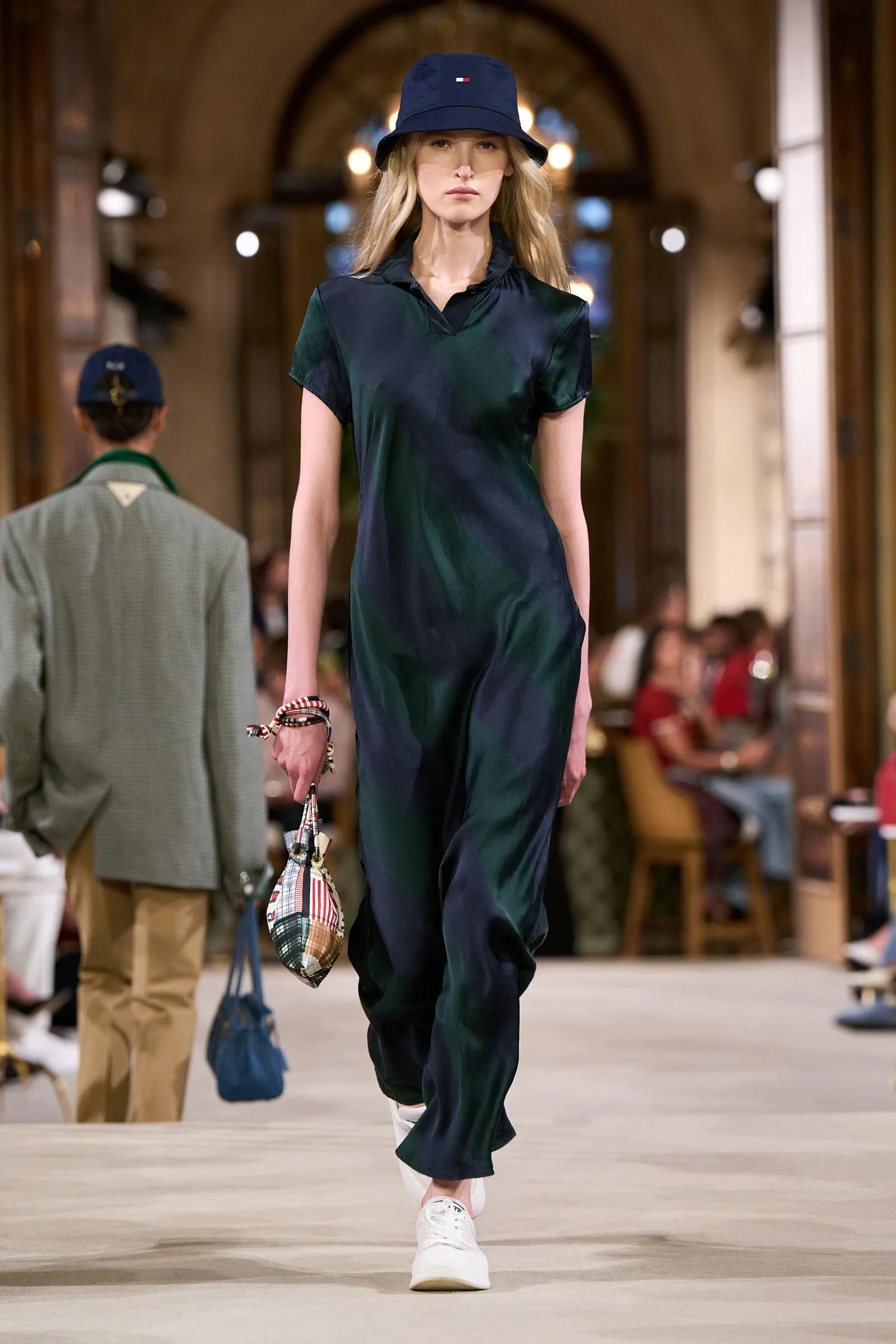 Female model walking a fashion runway in a dark green–navy satin jumpsuit and blue bucket hat, carrying a plaid patterned bag, inside a grand venue with onlookers in the background.