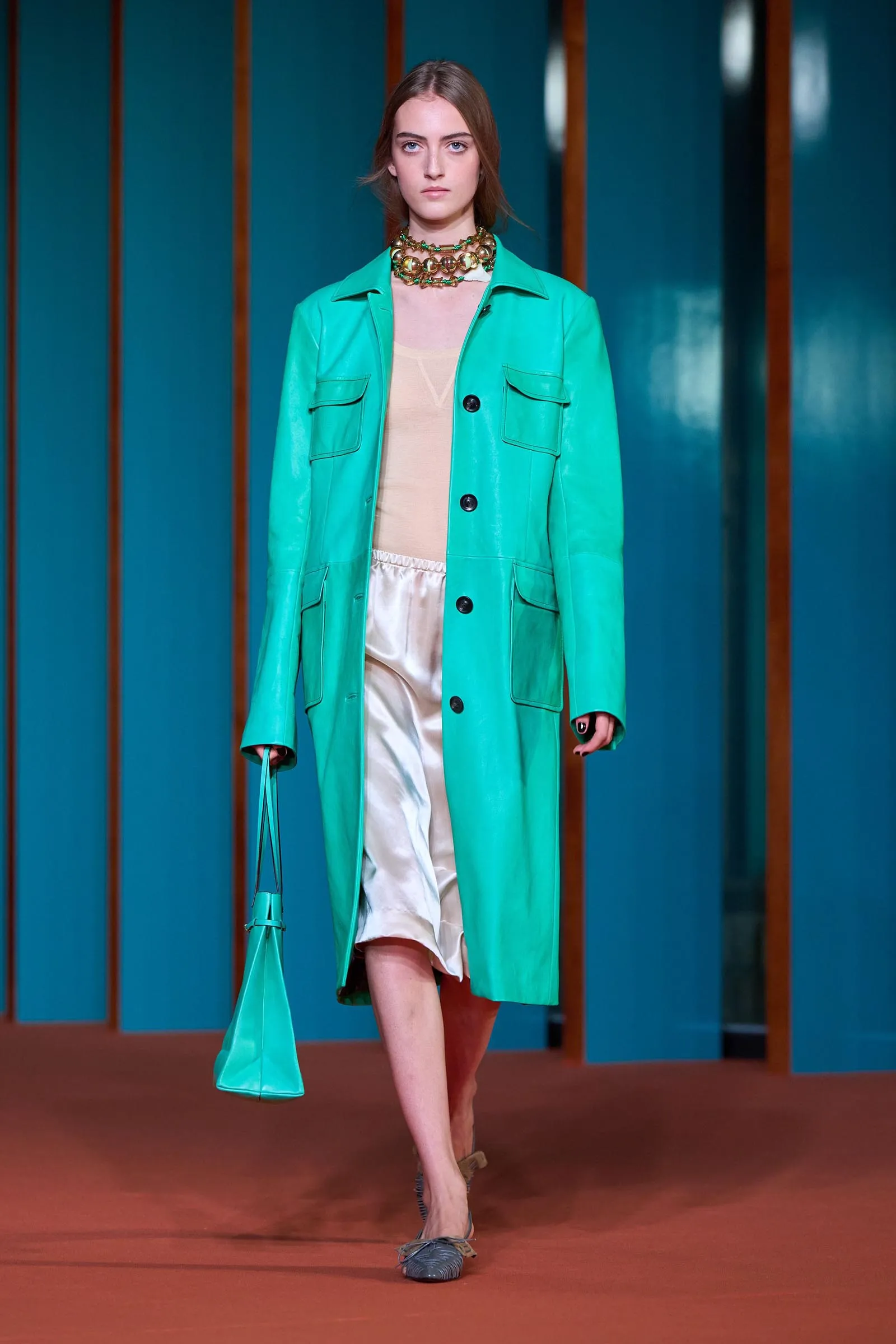 Model walking a runway in a bright teal coat over a beige top and cream satin skirt, with a chunky gold necklace and turquoise bag, against teal panels.