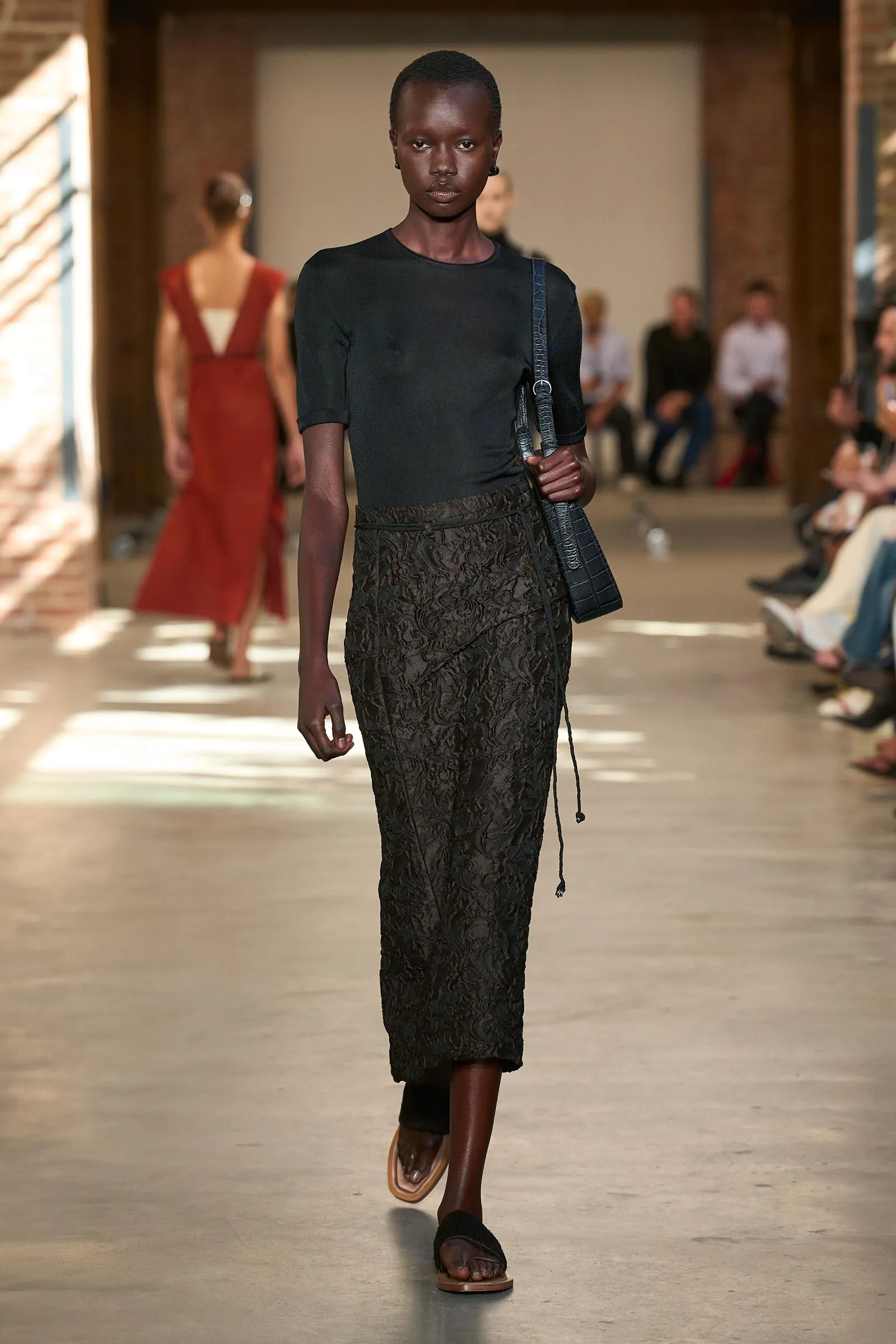 Runway model wearing a black top and dark patterned skirt, carrying a blue shoulder bag as she walks down the catwalk.