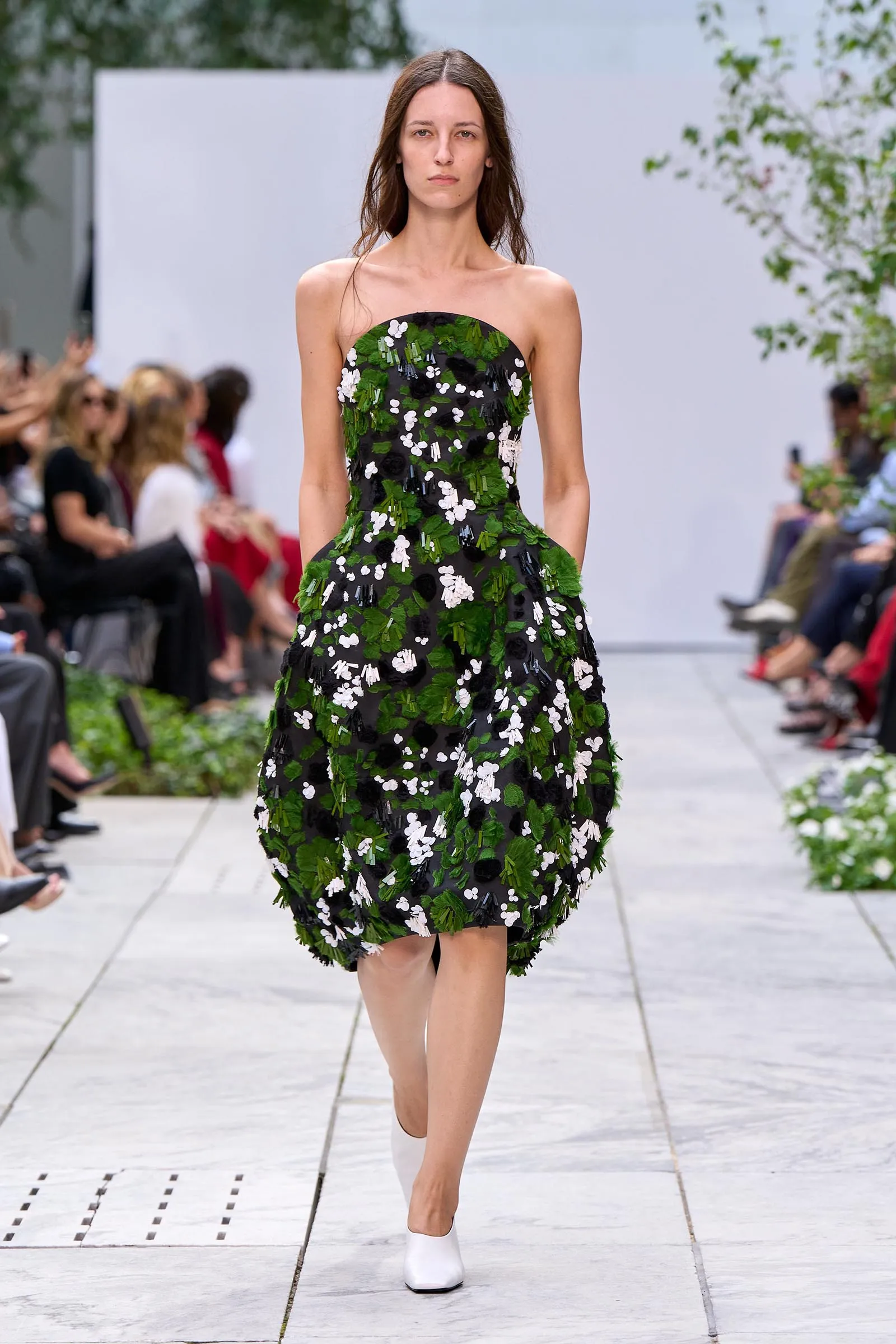 Model walks the runway in a strapless black dress adorned with green leaves and white floral appliqués.