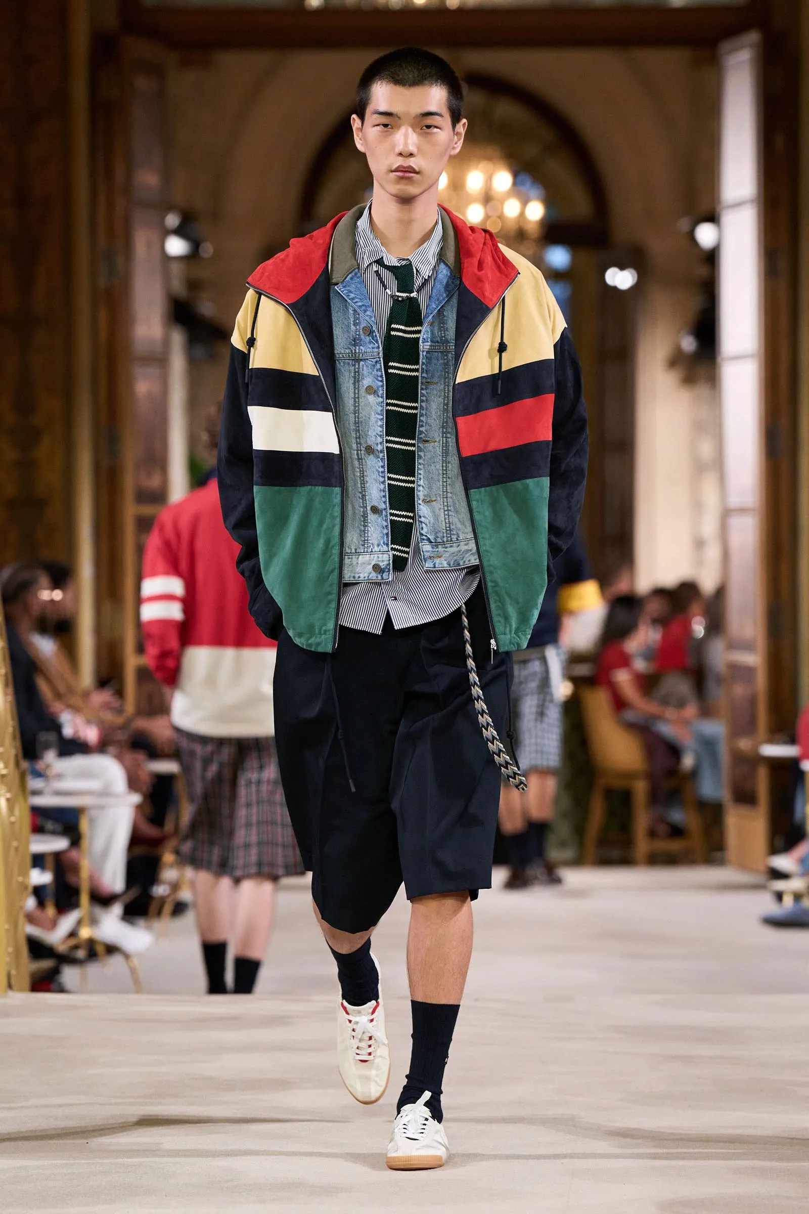 Male model walks a runway in a color-block jacket over denim layers, striped shirt, and tie; audience blurred in the background.