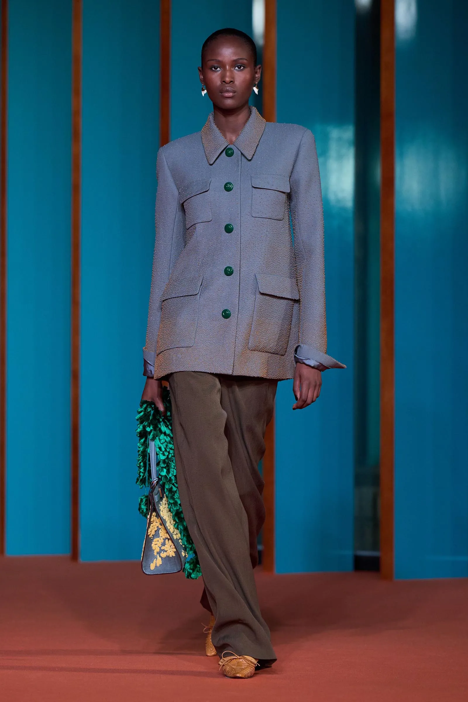 Model walking a fashion runway in a structured gray textured jacket with large flap pockets and green buttons, paired with brown pants and a patterned green accessory.