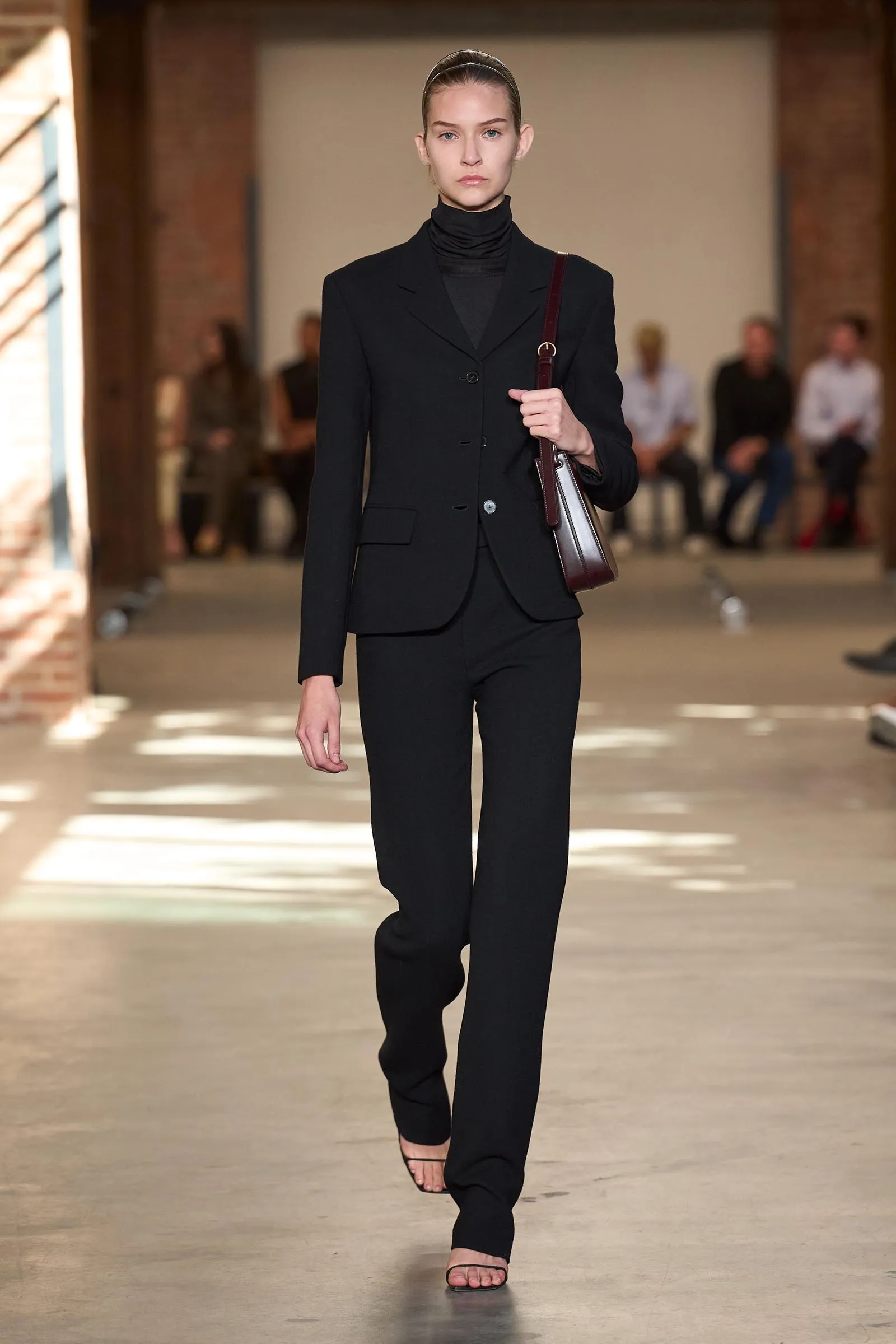 Female model walking a runway in a black tailored suit with a black turtleneck, carrying a burgundy handbag.
