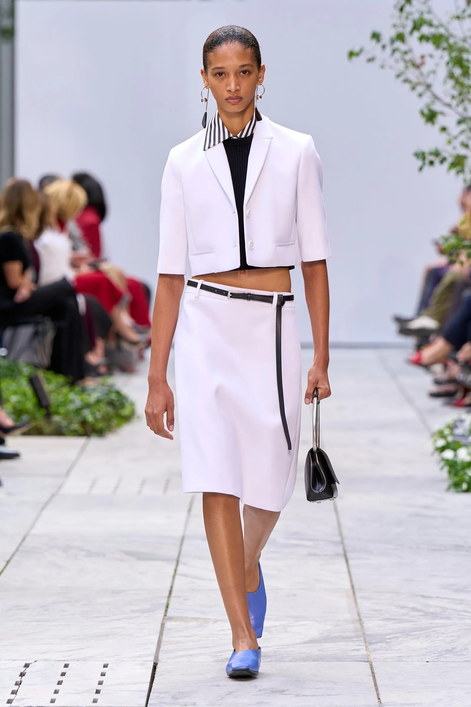 Model walking a runway in a white cropped blazer, black top, and white skirt, holding a black handbag; blue shoes visible.