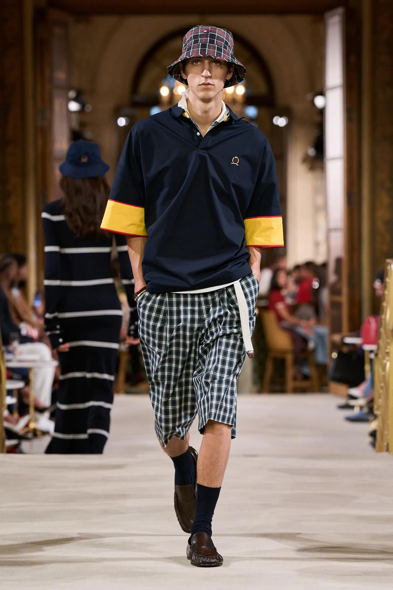 Male fashion model walking a runway in a navy polo with yellow-trimmed sleeves, plaid shorts, and a plaid bucket hat.