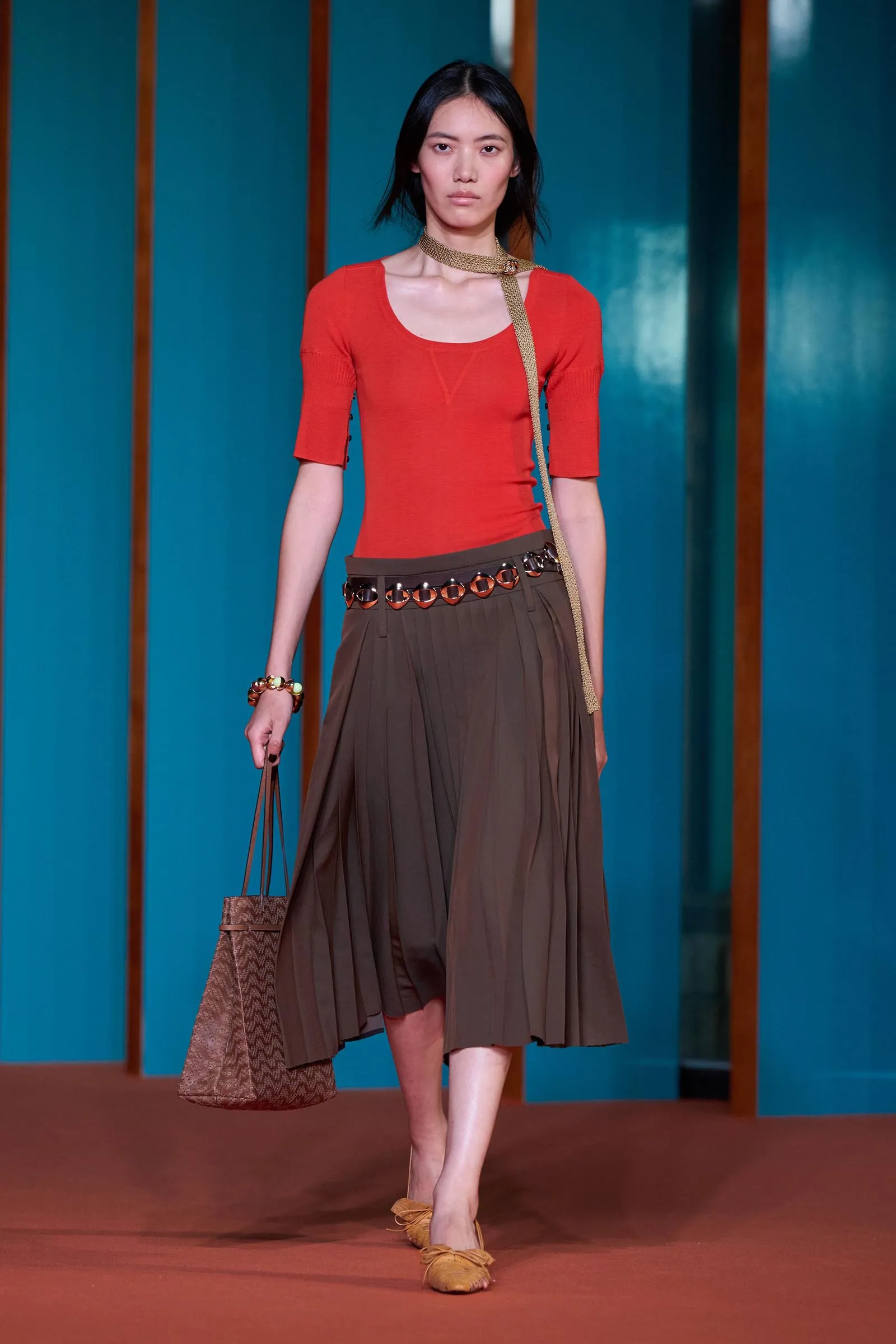 Model walking a runway in a red-orange top, brown pleated skirt, and gold accessories against a teal backdrop.