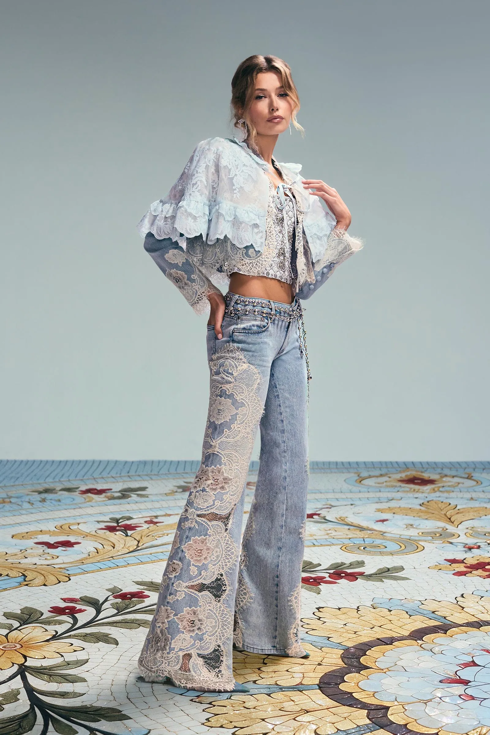 Woman posing in a light blue lace cropped top and embellished jeans, standing on a colorful floral tiled floor against a pale blue backdrop, fashion portrait.