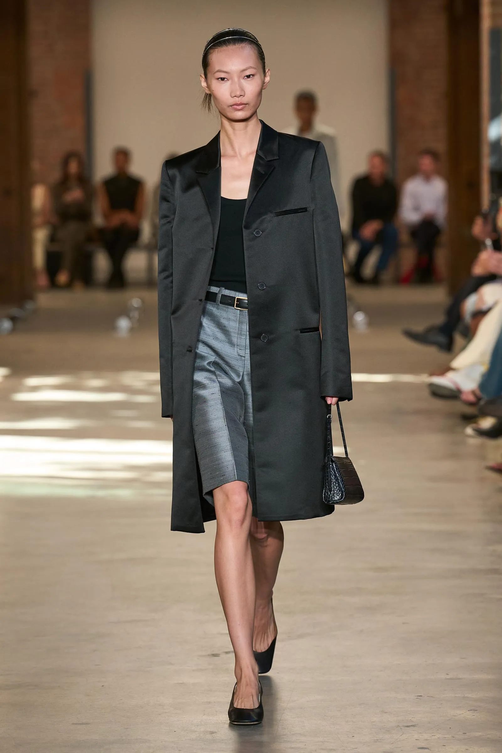 Model walking runway in a black blazer over a black top and gray satin skirt, carrying a small black handbag.