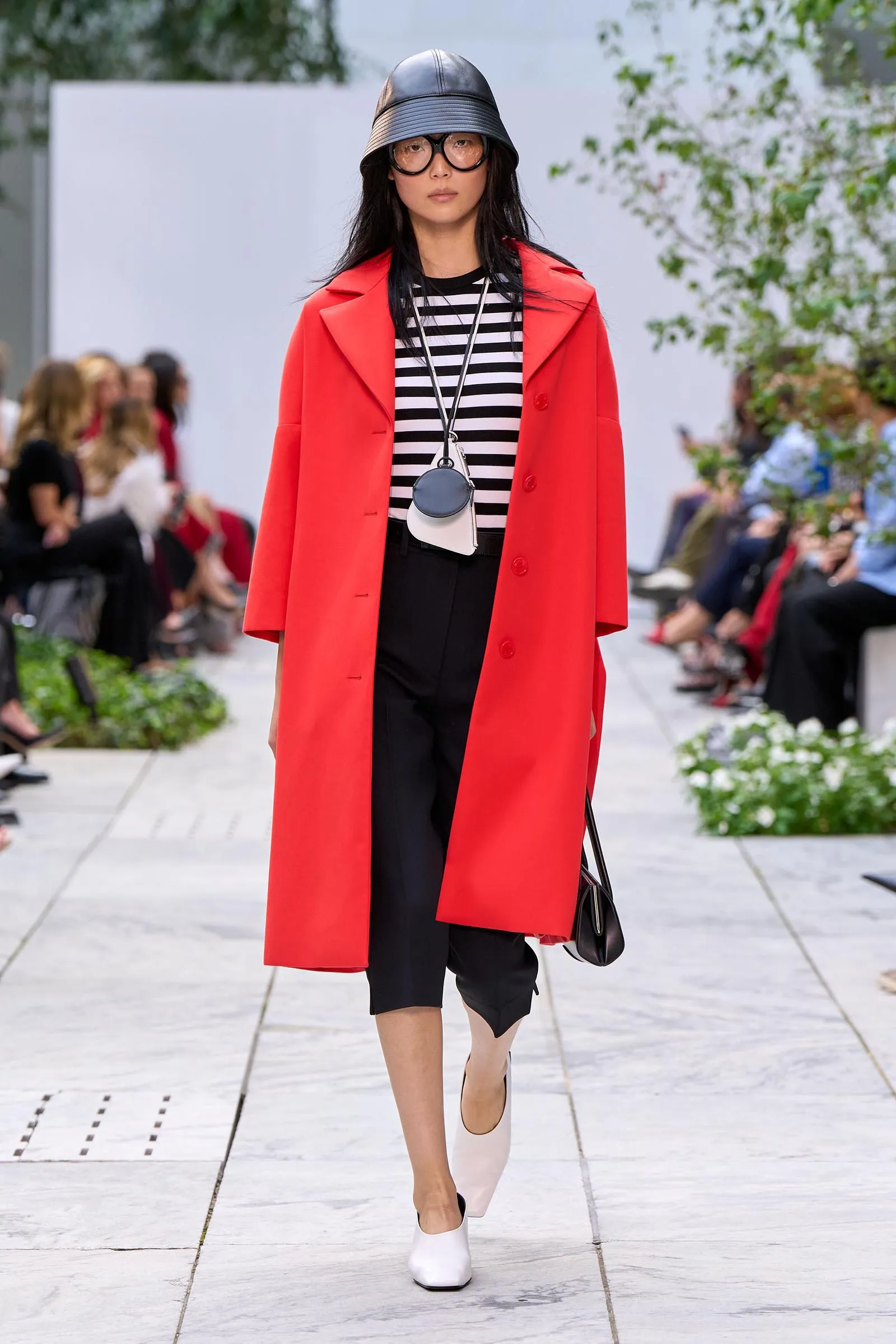 Model walking a fashion runway in a bright red overcoat over a black-and-white striped top, black cropped pants, white shoes, and a black bucket hat with oversized round glasses, carrying a small round bag.