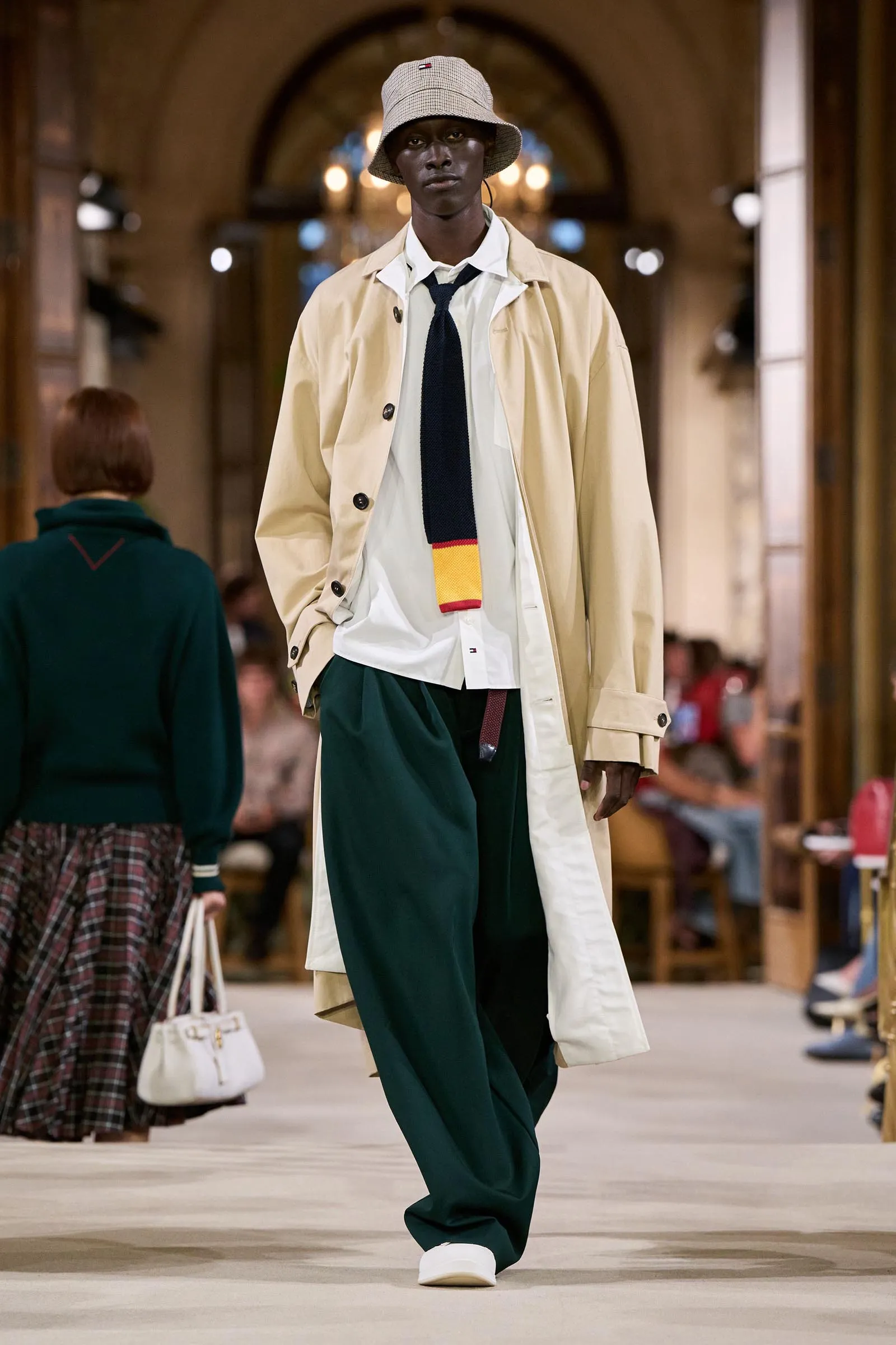 Model on a runway wearing a beige trench coat over a white shirt and dark tie, paired with green trousers and white sneakers, in a chandelier-lit hall with a bucket hat.