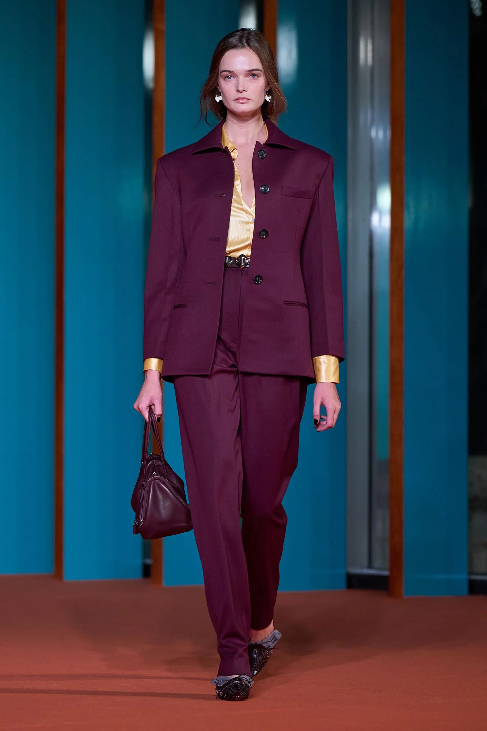 Model wearing a burgundy pantsuit with a gold satin blouse, carrying a matching burgundy bag on a runway.