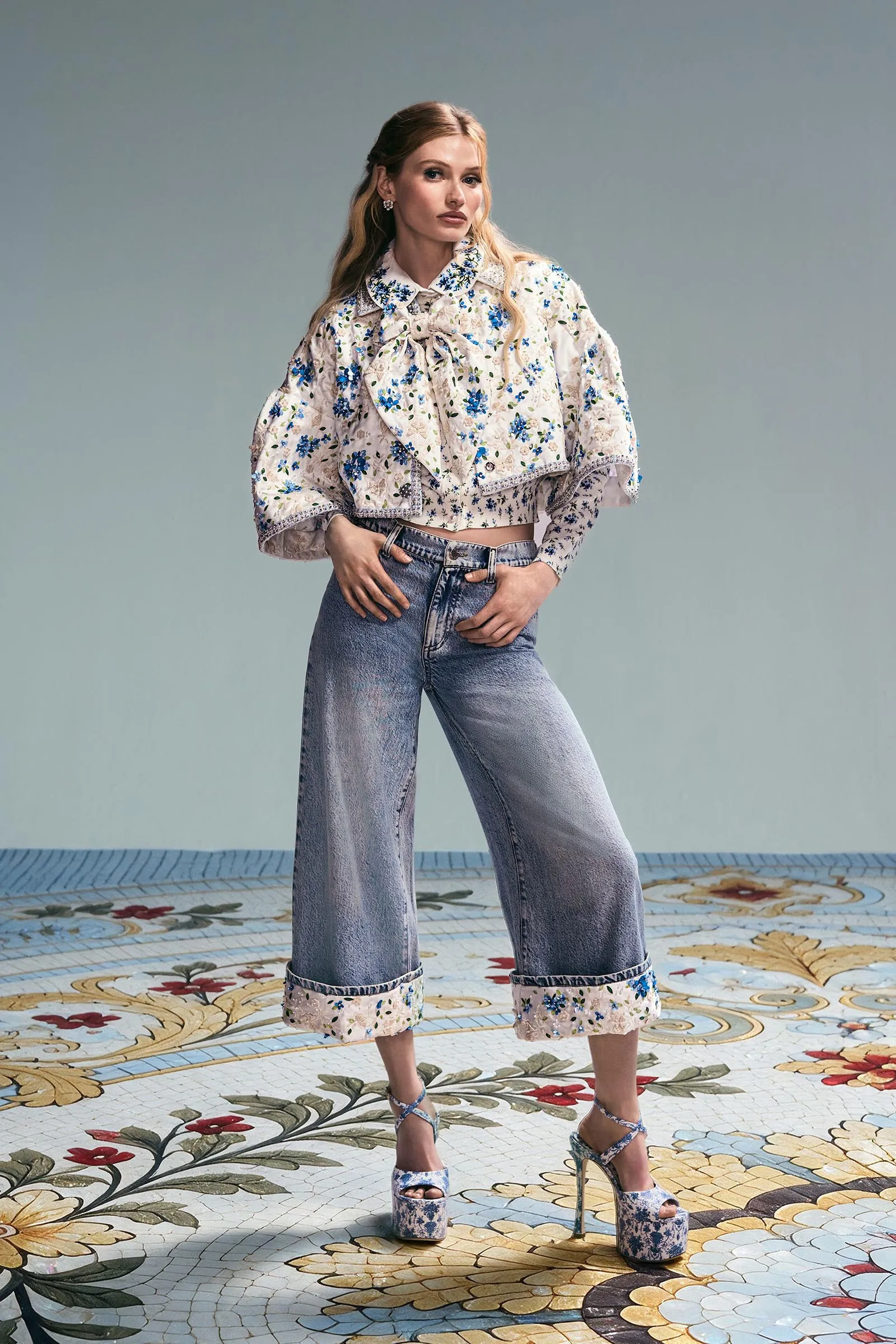 Model posing in a floral-pattern blouse and wide-leg jeans, hands in pockets, against a pale backdrop.
