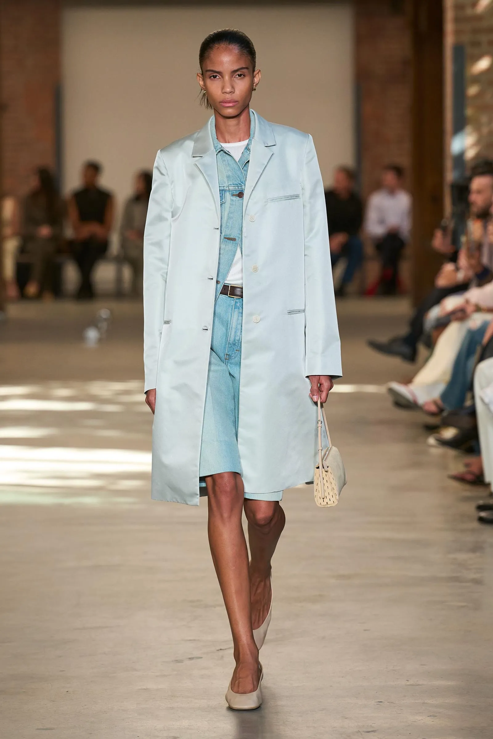 Model walks the runway in a pale blue oversized coat over a light denim outfit, carrying a small beige handbag.