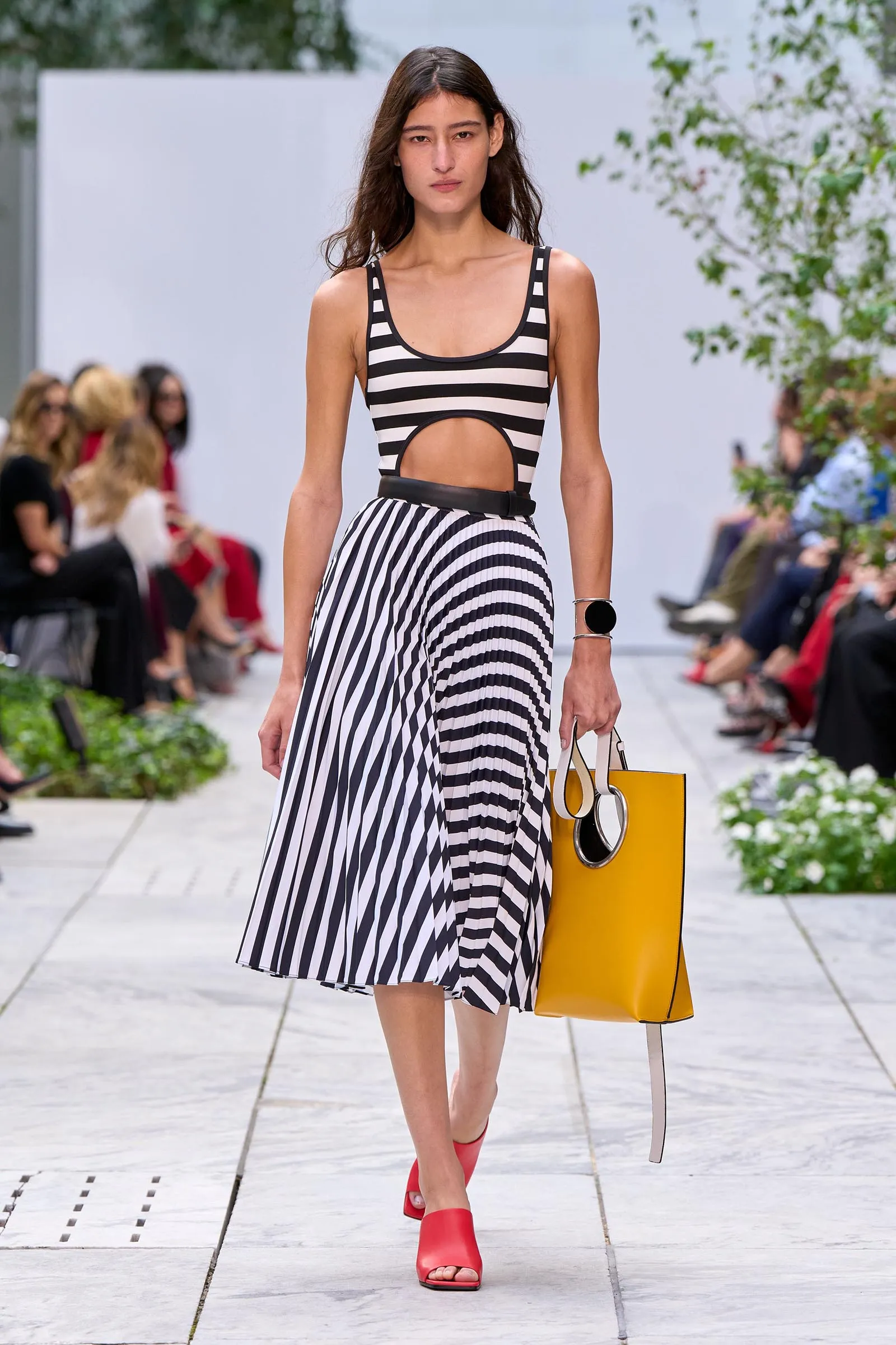 Model walks a fashion runway in a black-and-white striped sleeveless dress with a waist cut-out, carrying a yellow shopping bag and wearing red sandals.