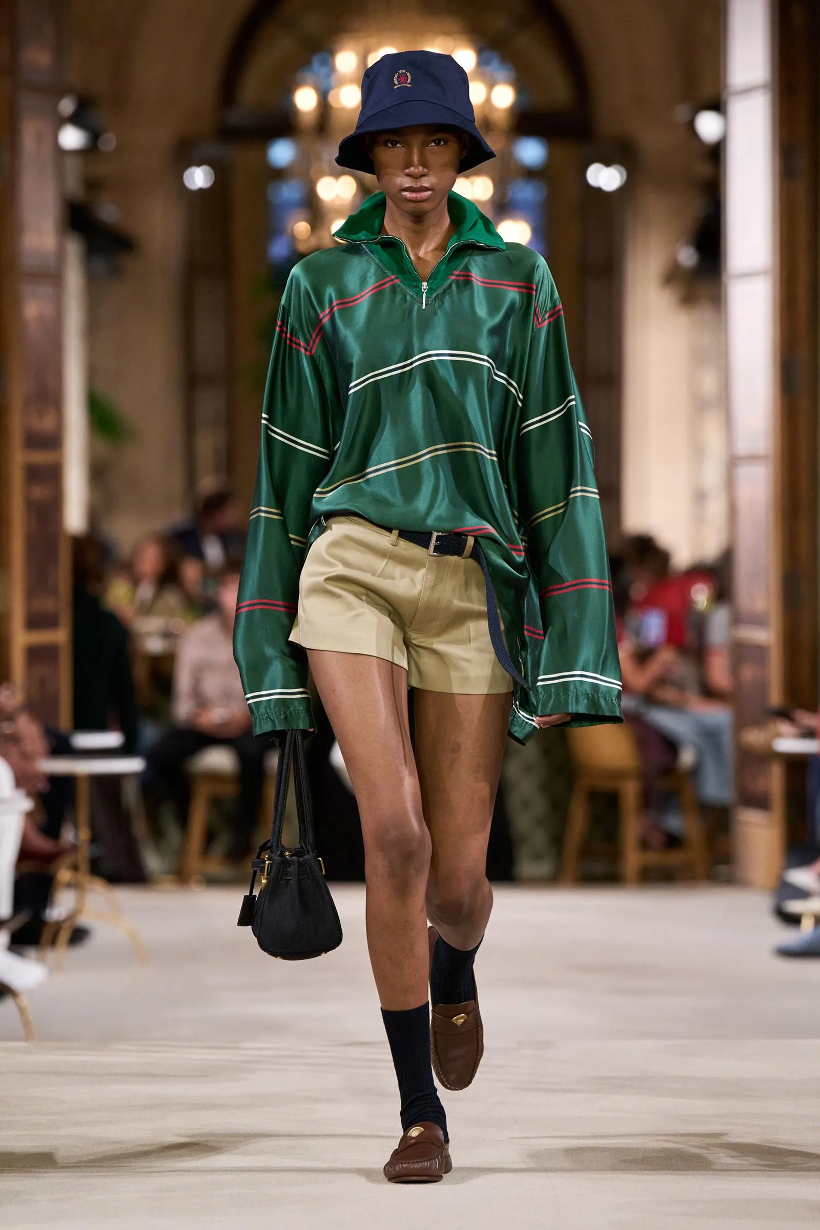 Model walking a runway in a green striped oversized top, beige shorts, and a navy bucket hat while carrying a black bag.