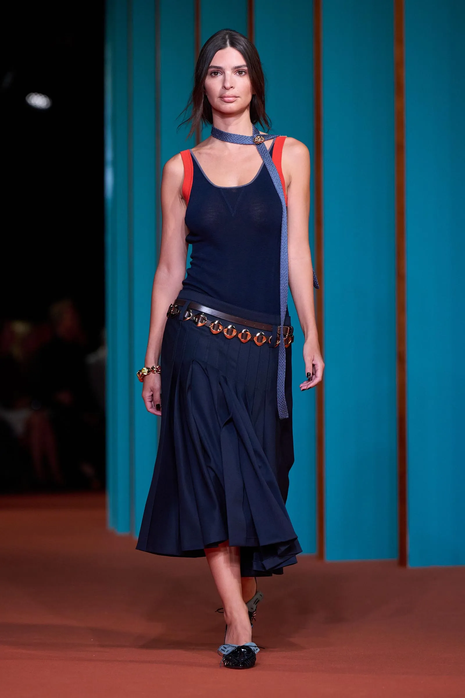 Model walking a runway in a navy sleeveless dress with red shoulder straps, a blue belt/scarf, and matching shoes against teal vertical backdrop.