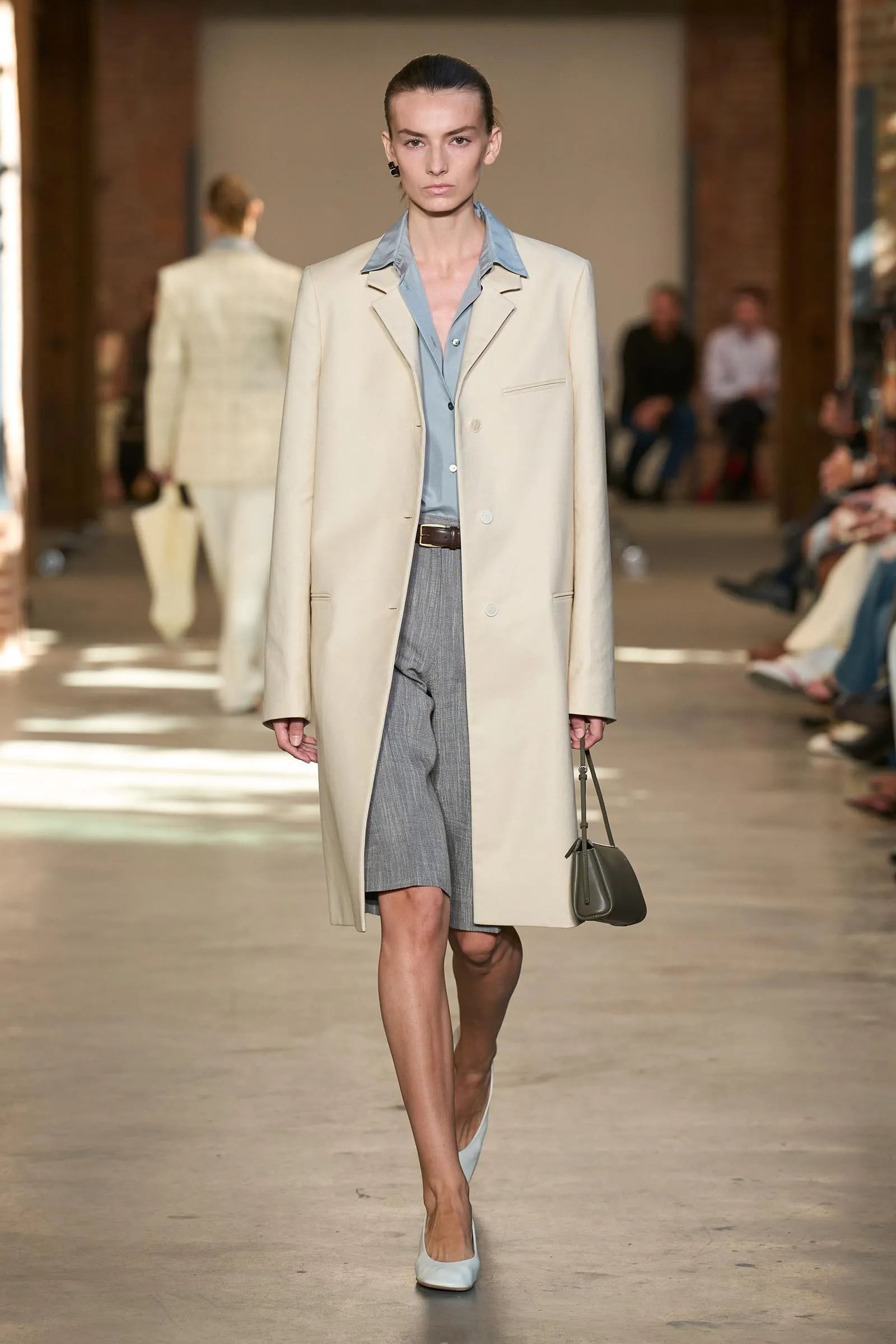 Model walking a runway in a cream overcoat over a light blue shirt, gray trousers, and light-blue heels, carrying a small gray handbag.