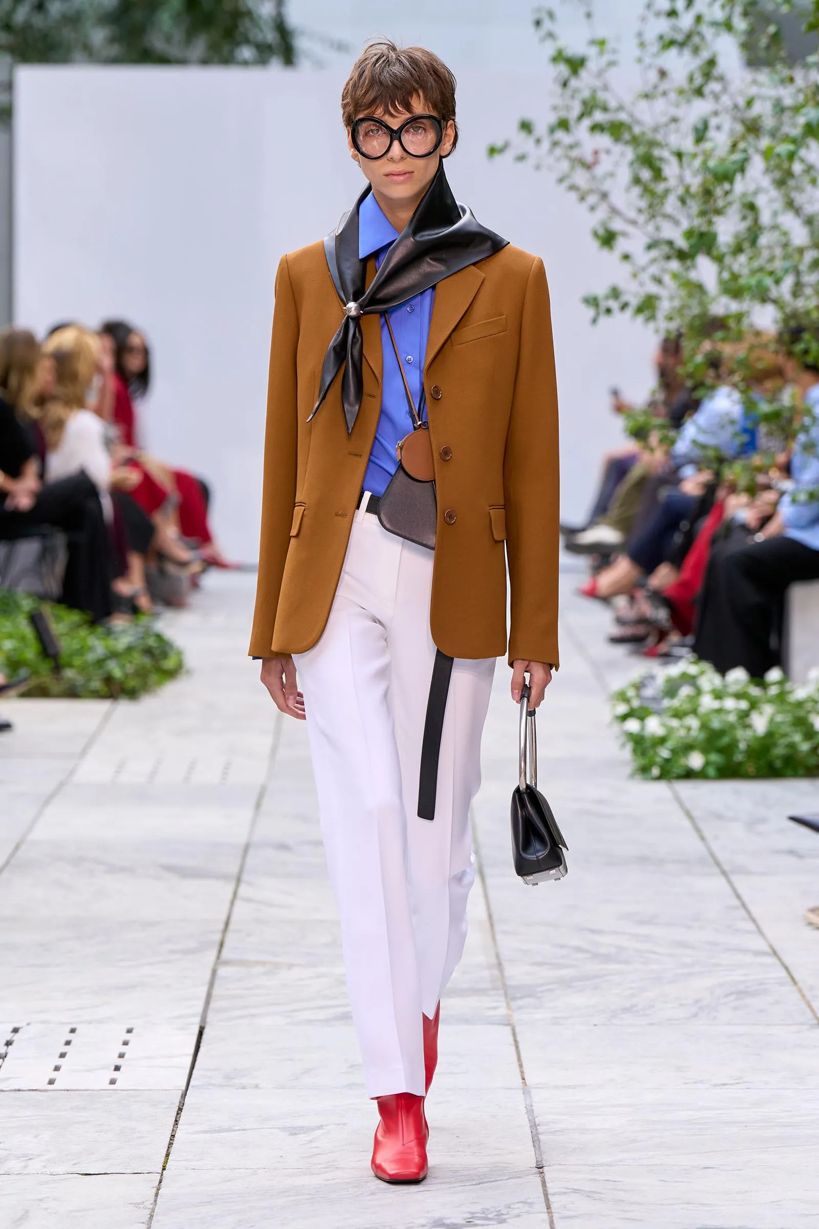 Model on a runway in a camel blazer over a blue shirt, white pants, red boots, a black scarf, and oversized round glasses.