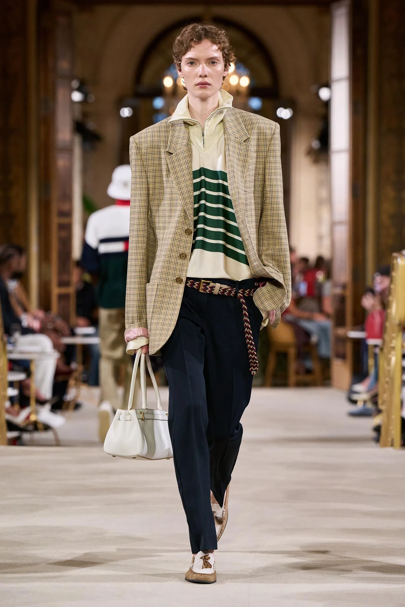 Model walks a fashion runway wearing a beige plaid blazer over a cream and green striped top, dark trousers, and a white handbag.