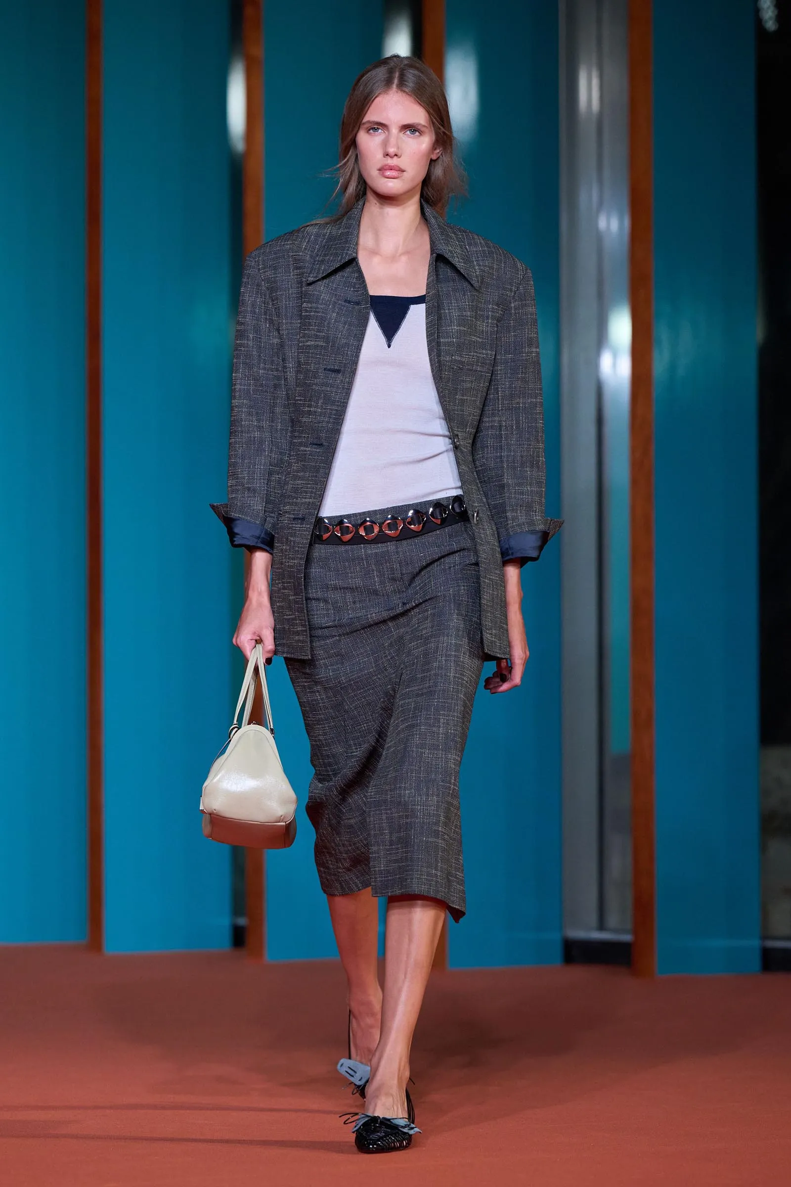 Model walking a runway in a gray plaid blazer and matching trousers, white top, and a light-colored handbag at her side.