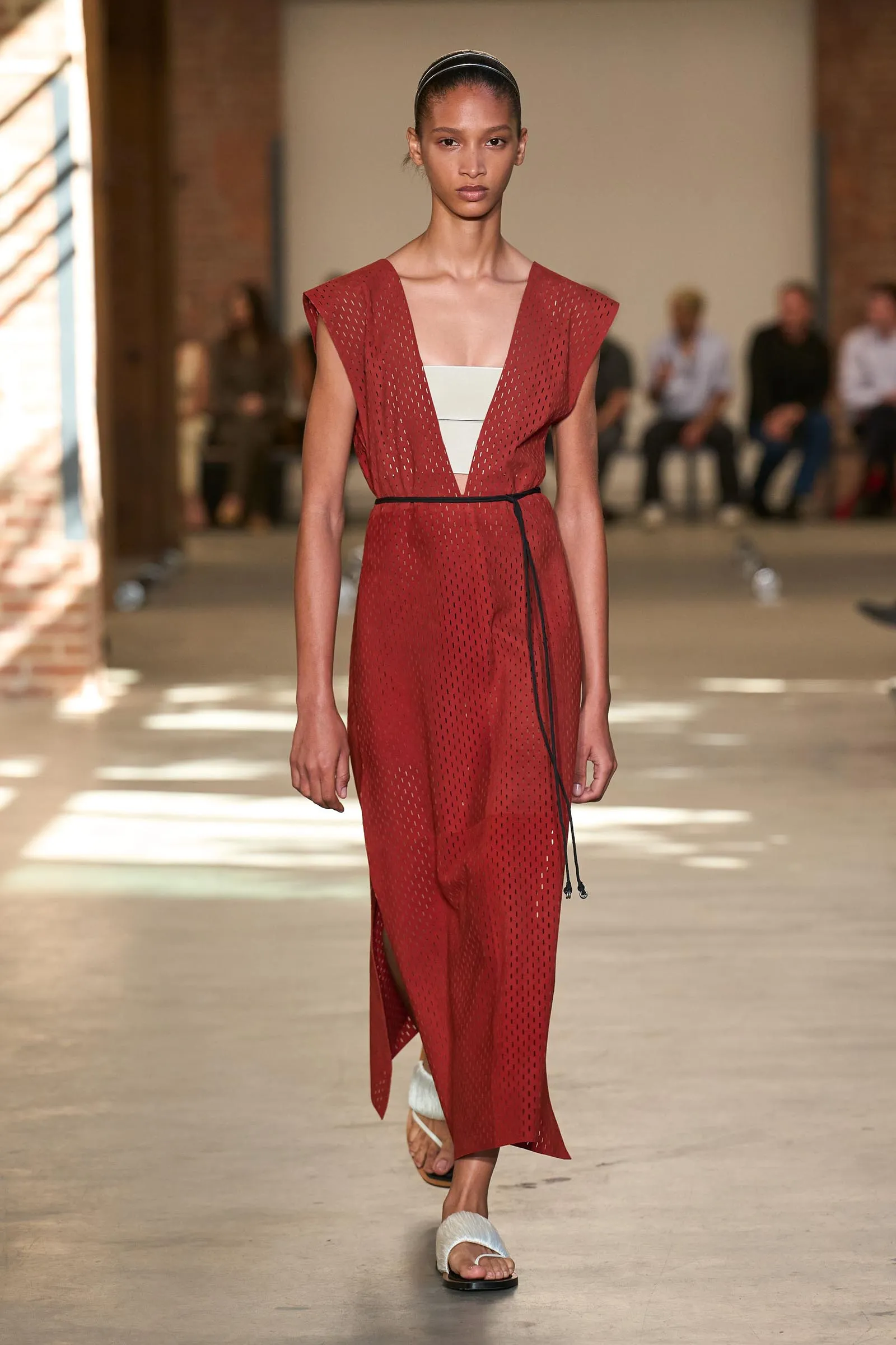 Model walks a runway wearing a red perforated sleeveless dress with a white inner panel and a thin black belt, white headband, and white sandals.