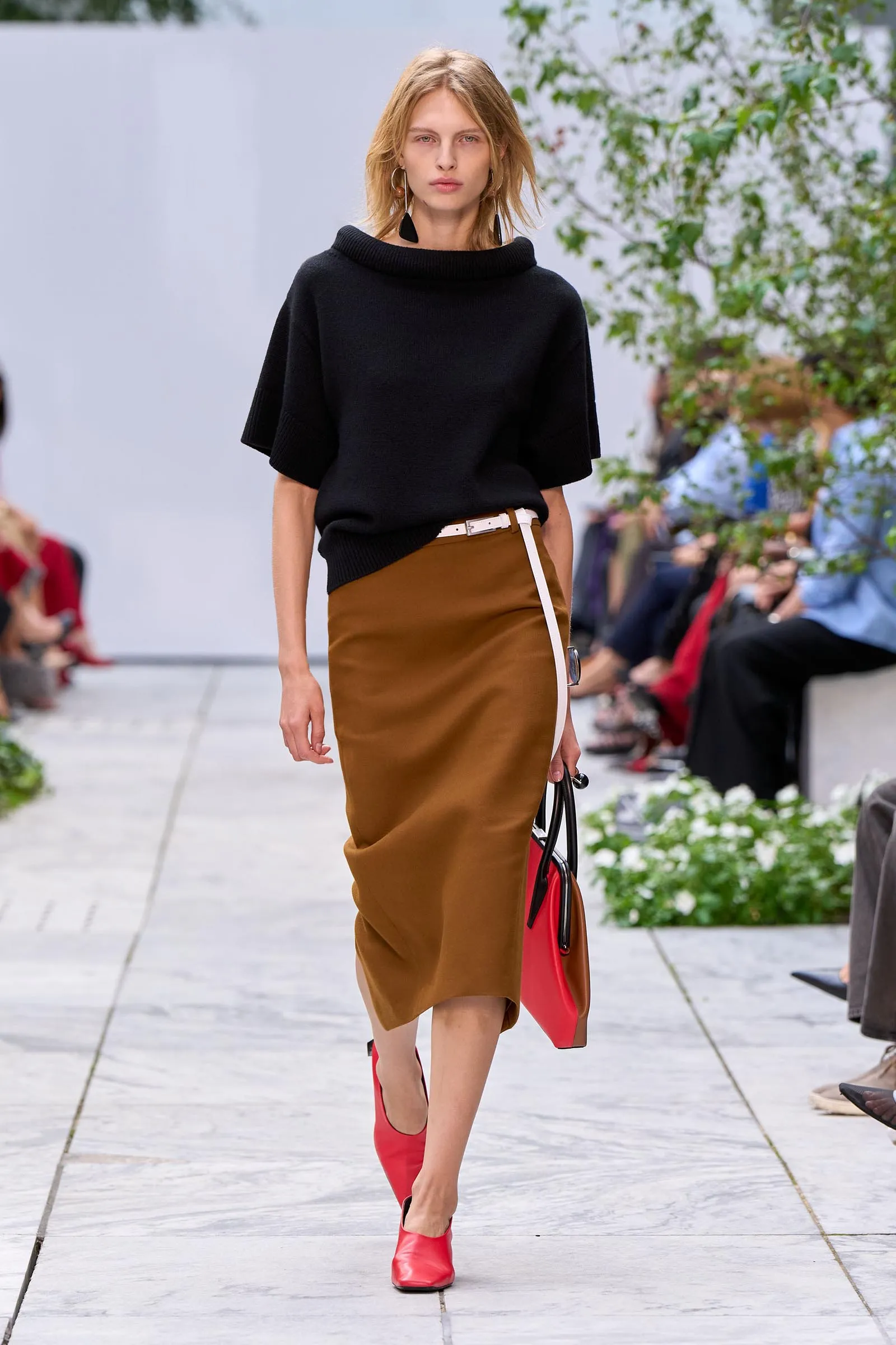 Model walking down a runway in a black oversized knit top, tan leather skirt, red ankle boots, and a red handbag.