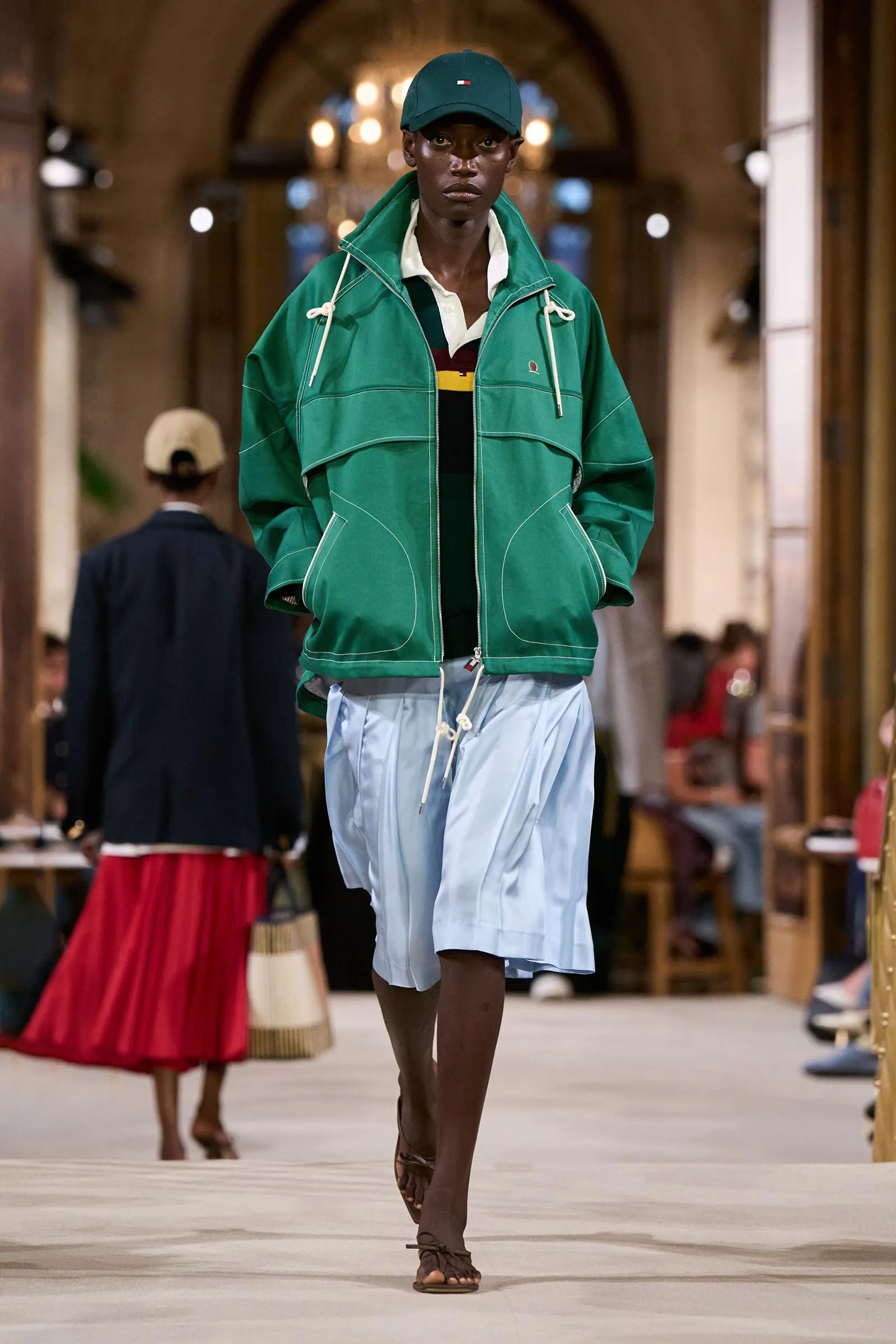Model walking a runway in a green windbreaker over a white-collar shirt and light blue shorts with a green cap and sandals under bright lighting.