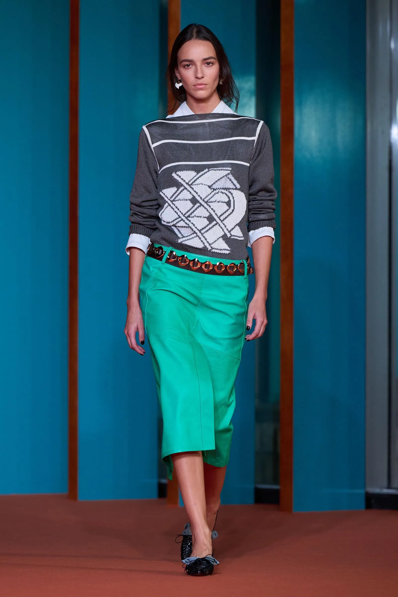 Female model walking a fashion runway in a gray sweater with white stripes and geometric design, paired with bright teal-green pants and black shoes.